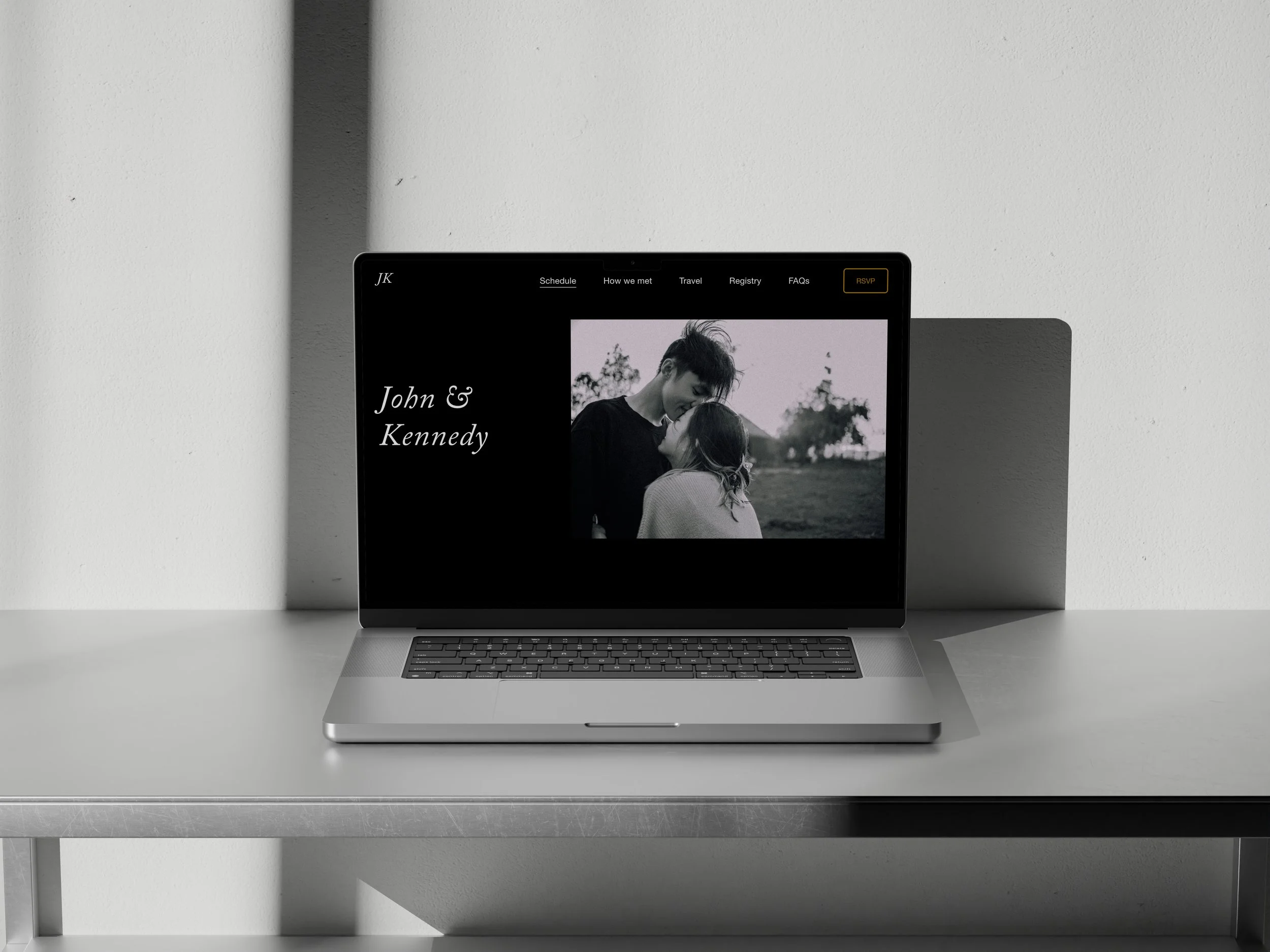 A silver laptop on a white table displaying a wedding website for John & Kennedy, with a black-and-white photo of a couple embracing outdoors.
