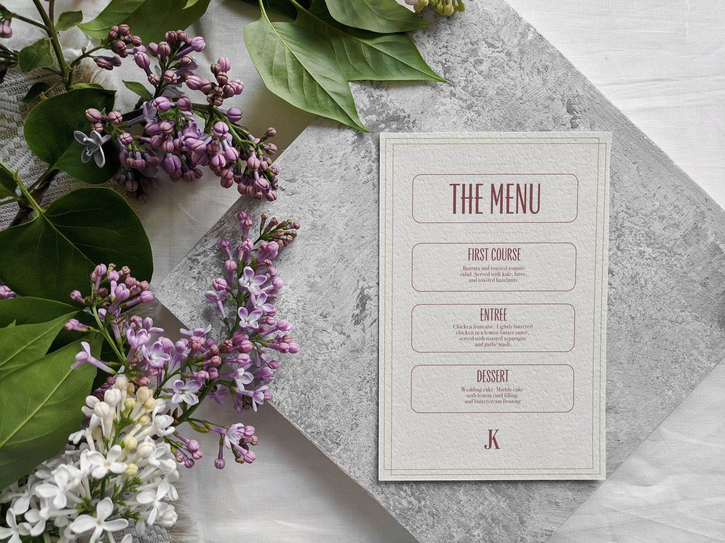 Menu card on gray stone surface accompanied by purple, white, and green flowers and leaves.