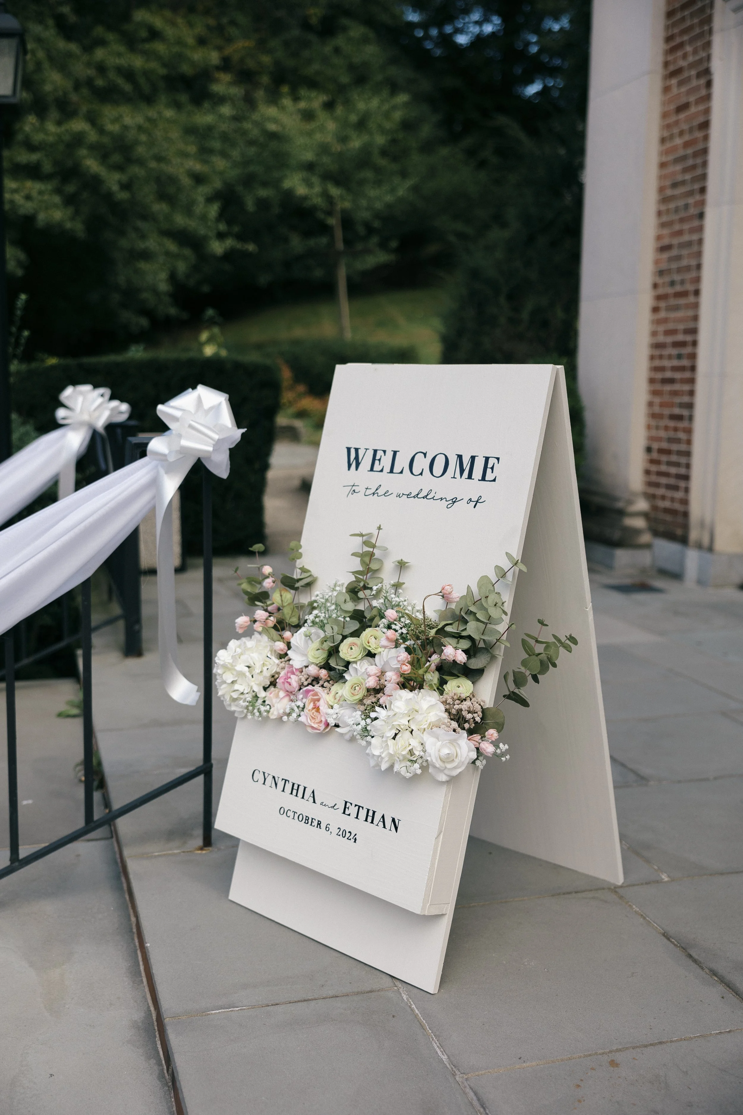 Wedding welcome sign with floral decorations on a sidewalk outside a building.