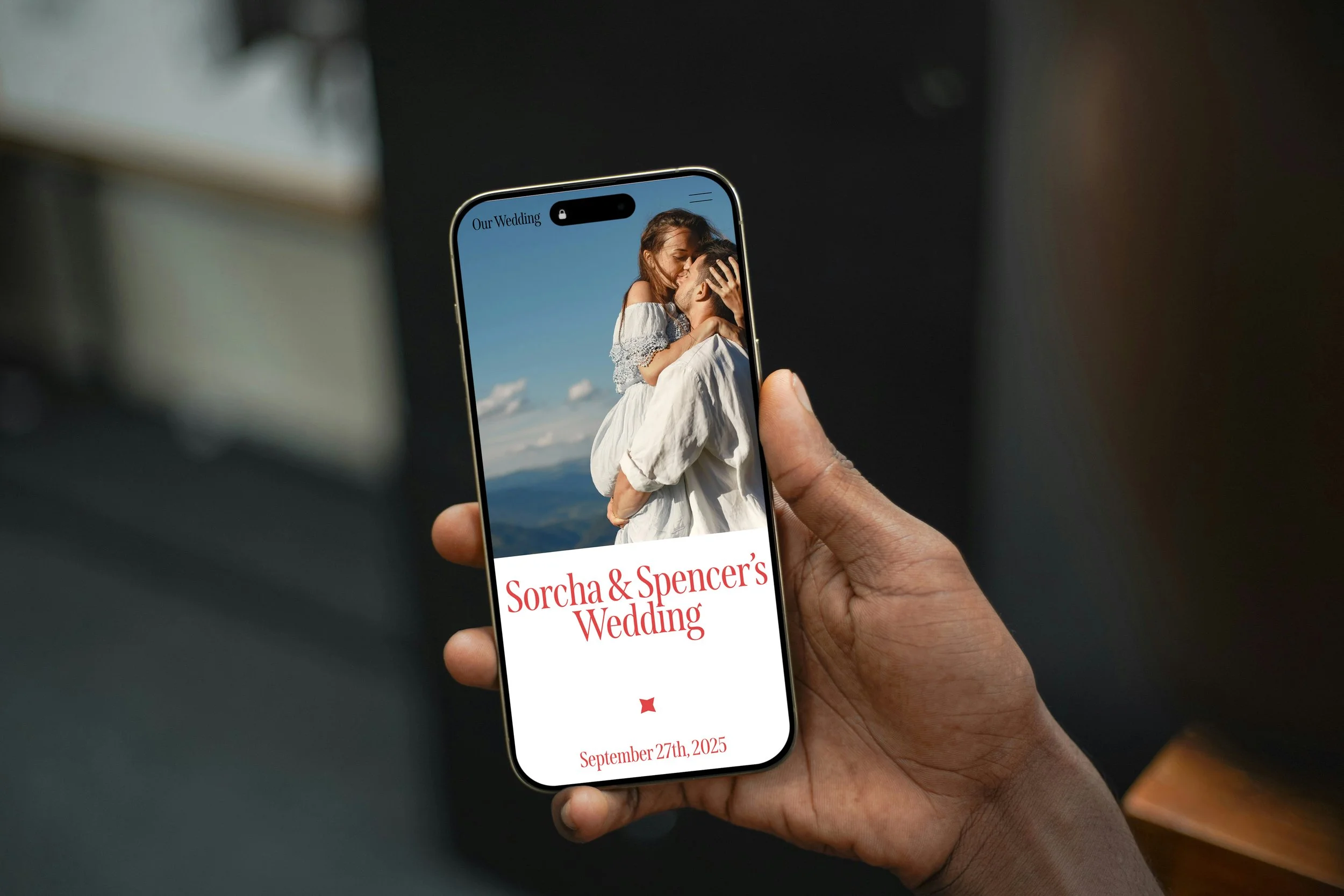 A person holding a smartphone displaying a wedding website for Sorcha and Spencer's wedding, featuring a photo of a woman and a man embracing against a mountainous backdrop.
