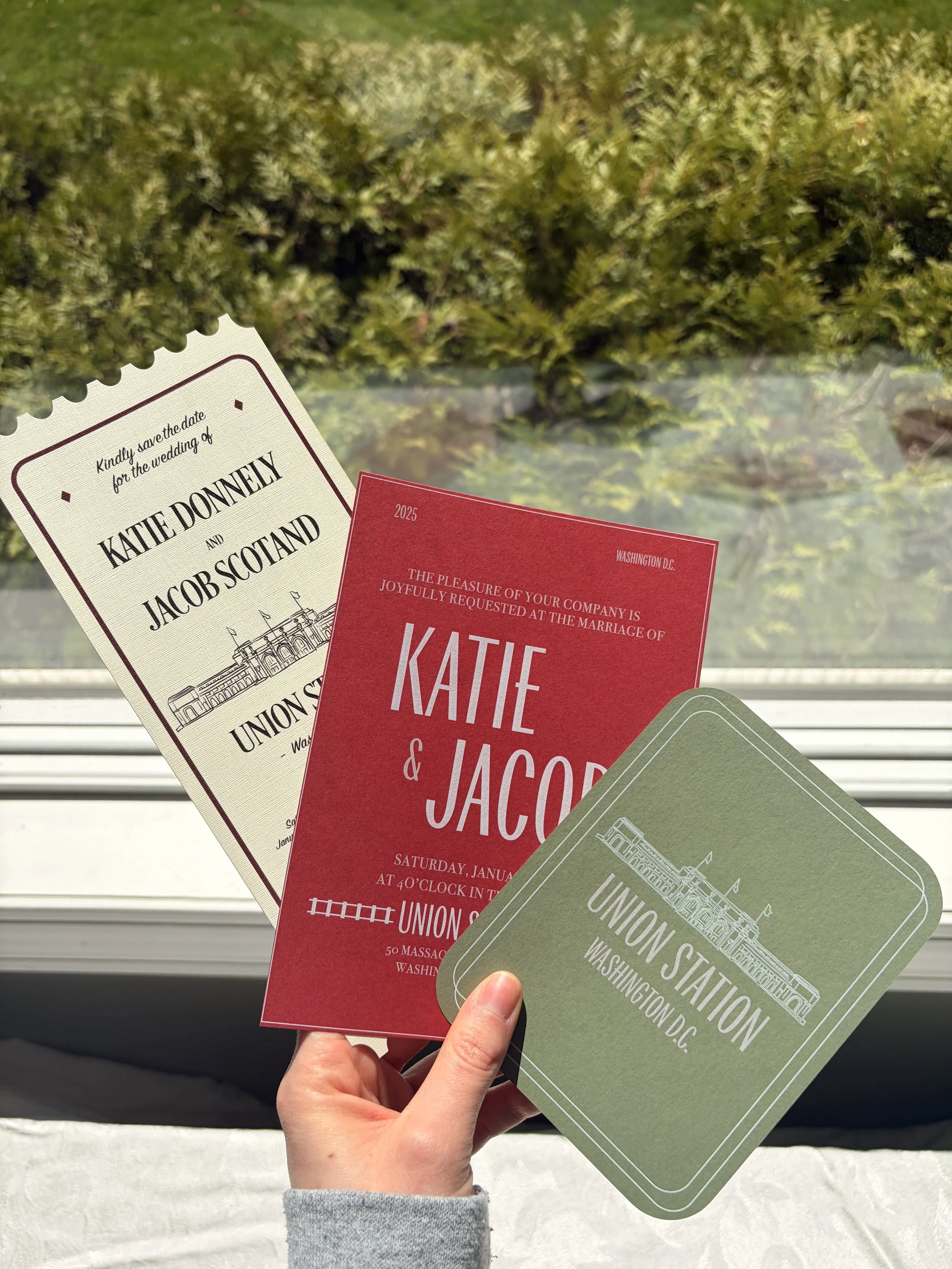 Hand holding three wedding invitations in front of a window with green shrubbery outside. One invitation is cream with black text, another is red with white text, and the third is green with white text and illustration.