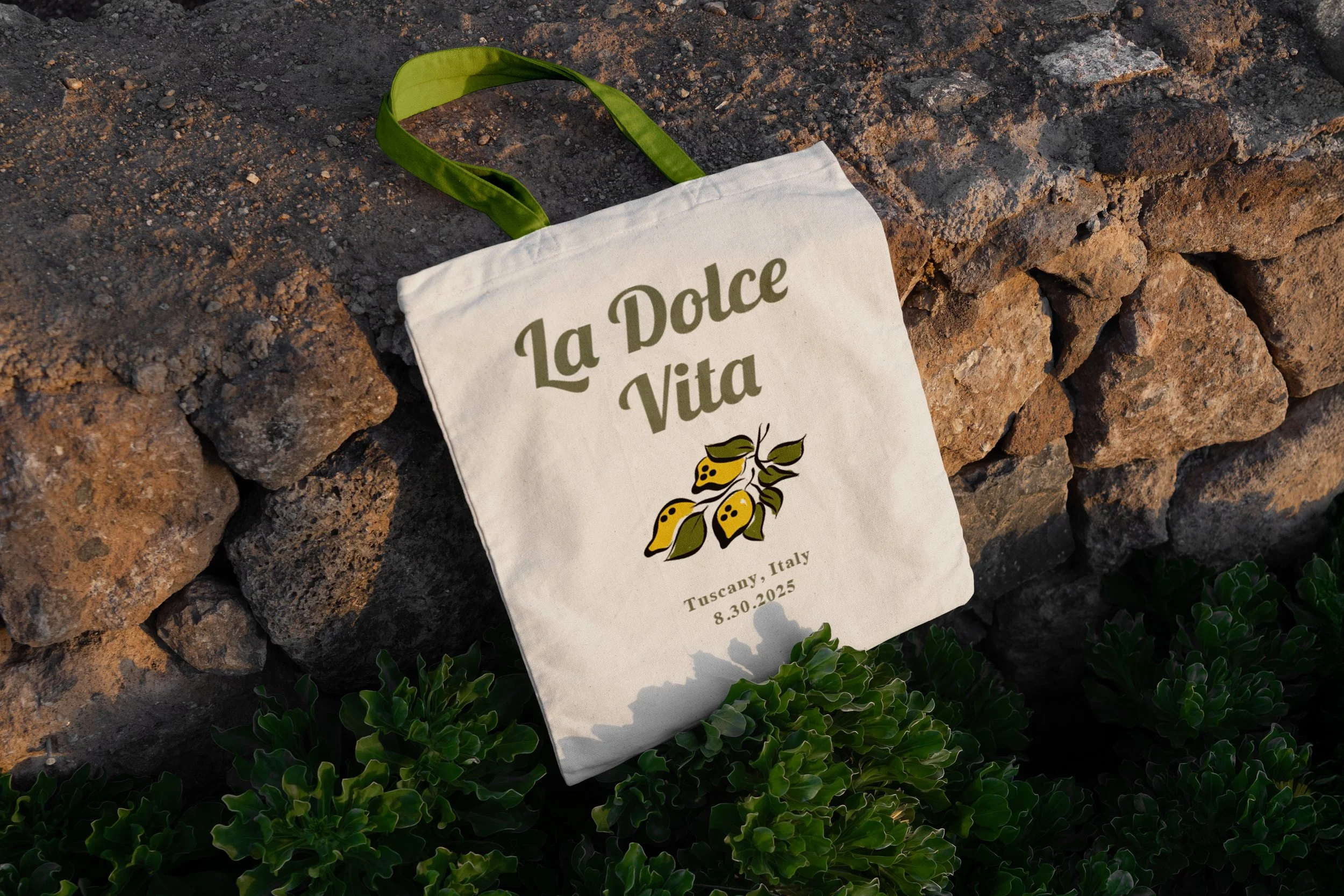White tote bag with green handles resting on a stone wall, displaying the words "La Dolce Vita," a lemon illustration, and the date August 30, 2025, in Tuscany, Italy, surrounded by green foliage.