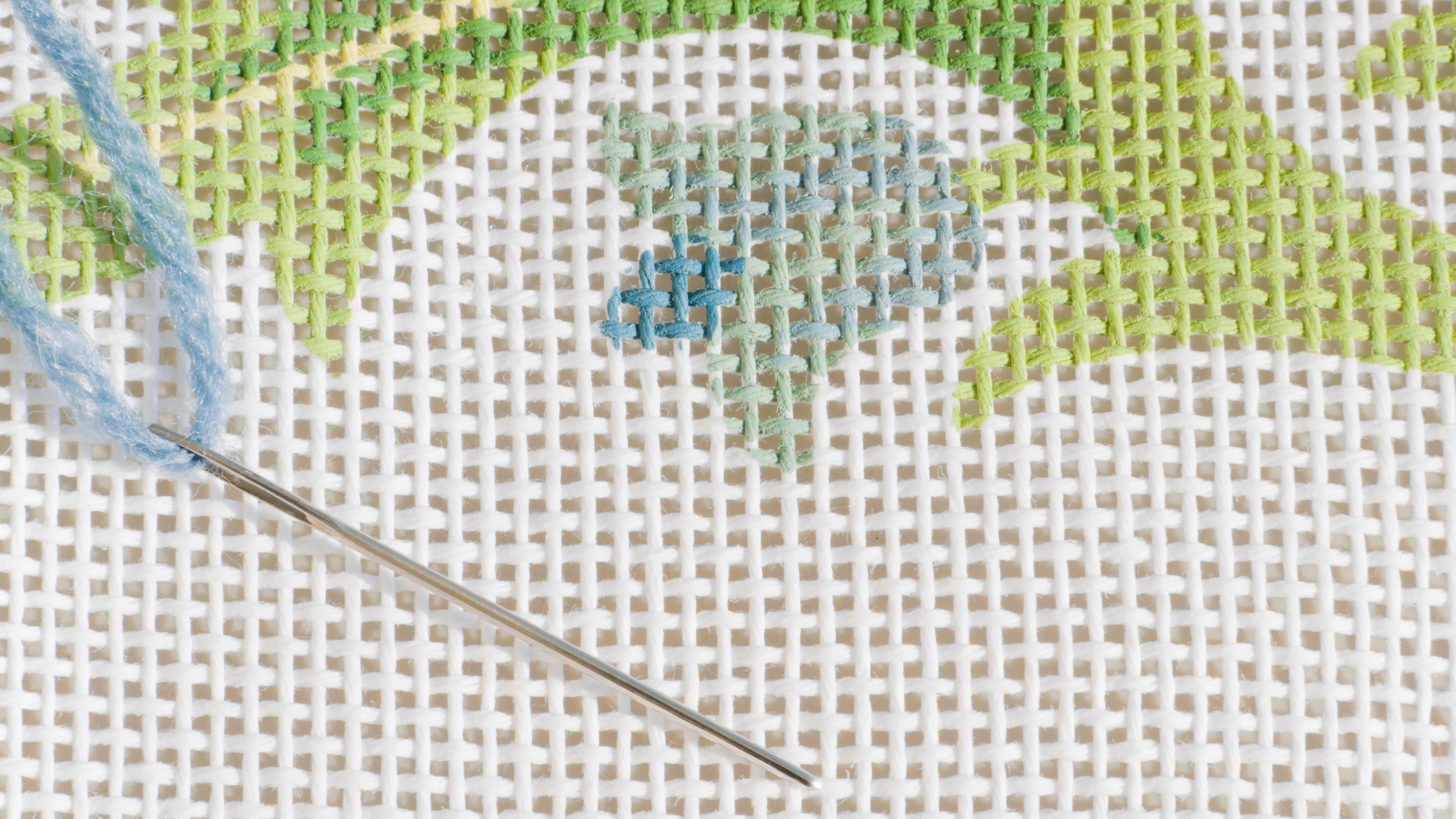 Close-up of embroidery cross-stitch fabric with a partially embroidered light blue and green design on white Aida cloth, with a sewing needle threaded with light blue thread resting on the fabric.