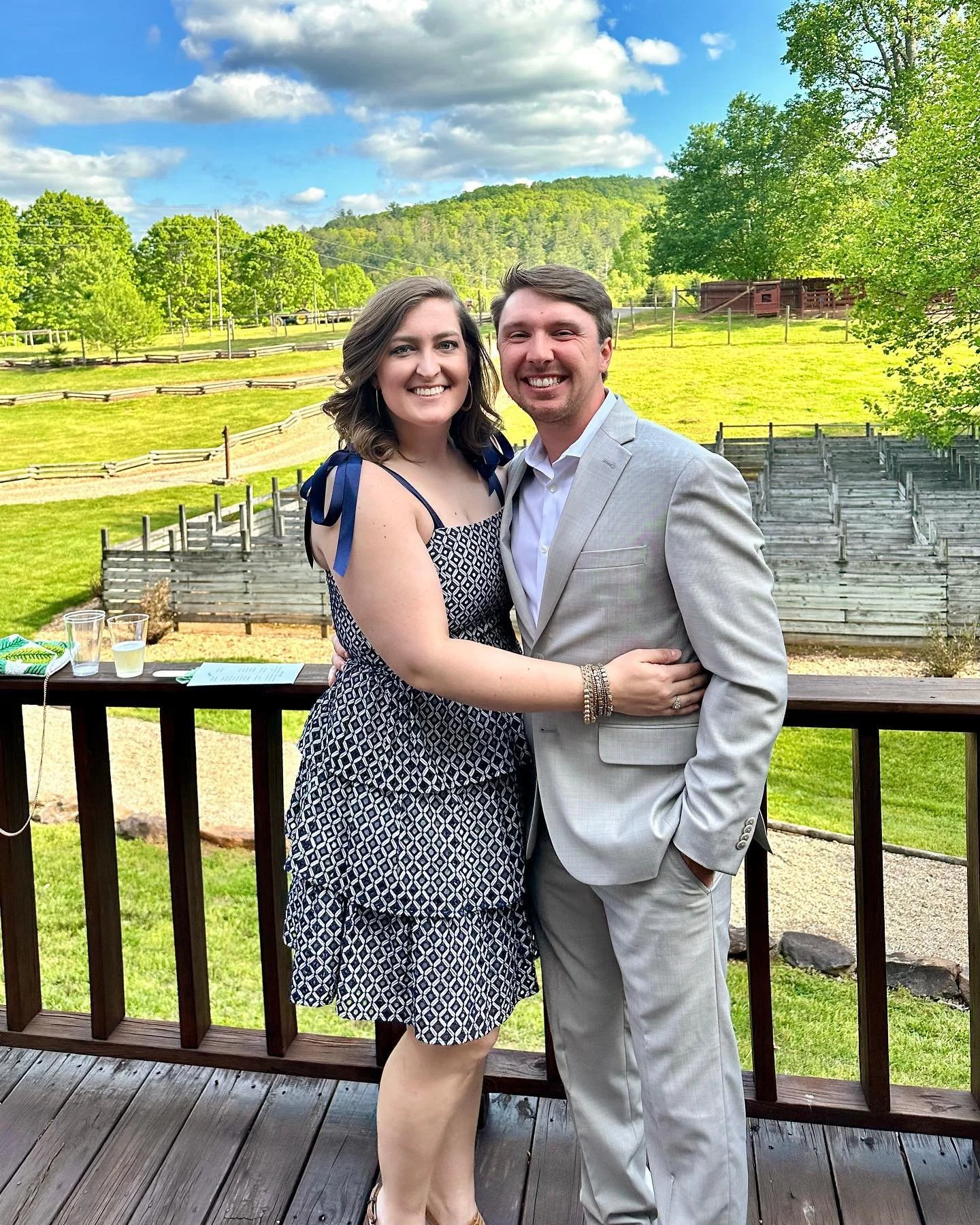 A smiling woman and man in formal attire standing close together outdoors on a wooden deck, with a scenic green landscape, trees, and hills in the background.
