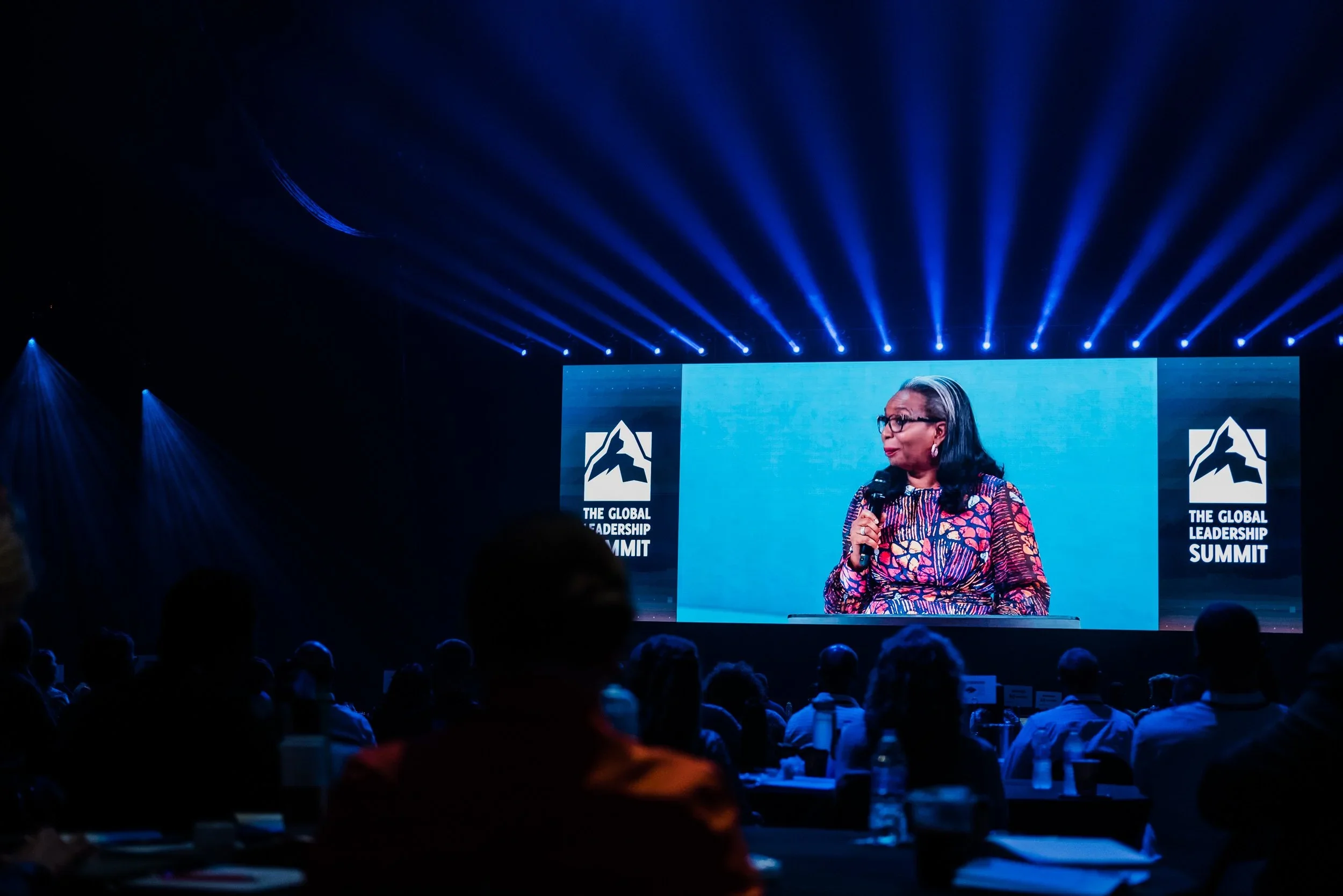 A woman speaking at a conference on stage with a large screen displaying the logo and text 'The Global Leadership Summit' in the background. The audience is seated in front with some silhouettes visible.
