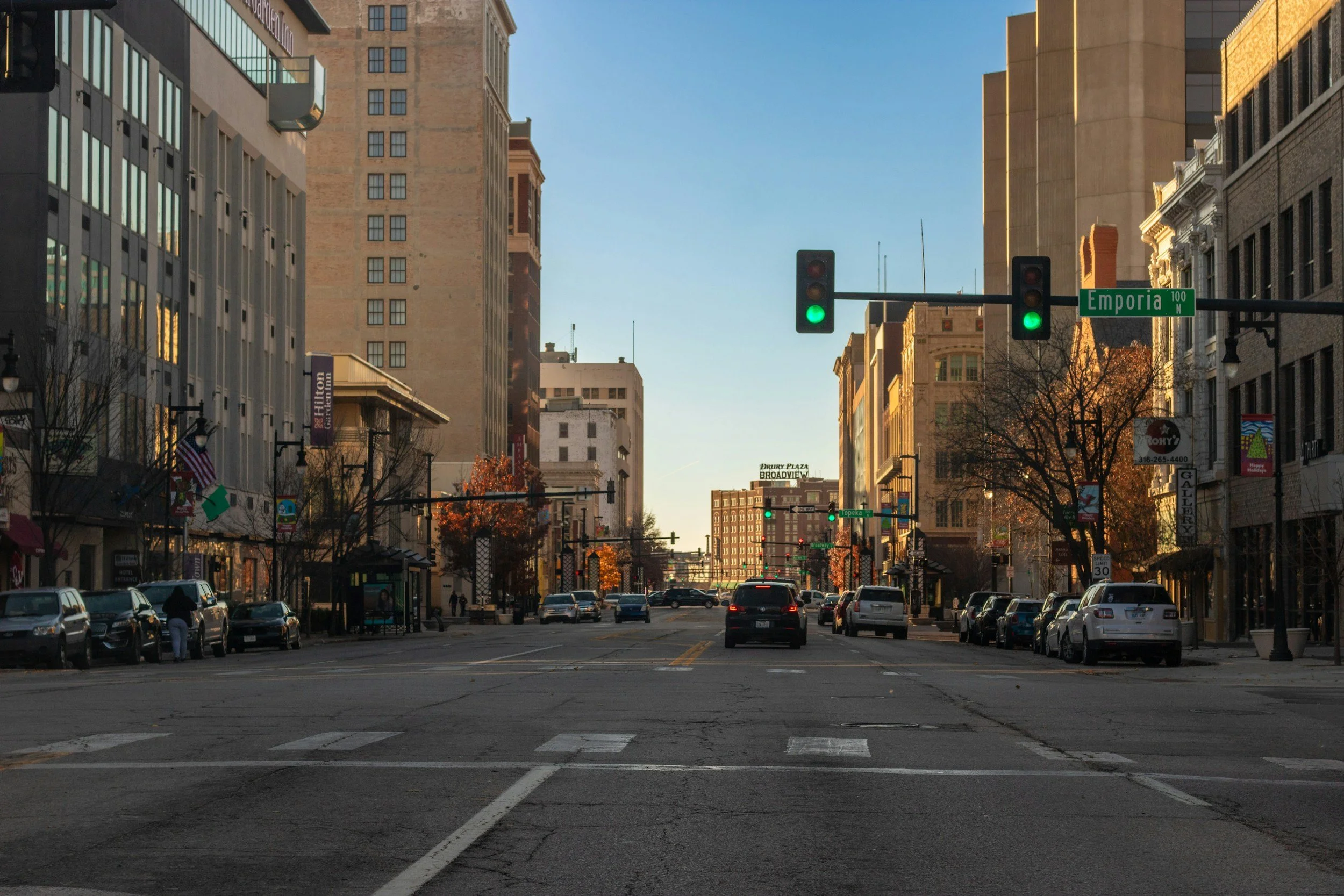 City street scene with traffic lights showing green, cars parked along the sides, tall buildings, and a clear blue sky.