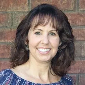 A woman with shoulder-length dark brown curly hair smiling in front of a brick wall, wearing a blue patterned top and earrings.