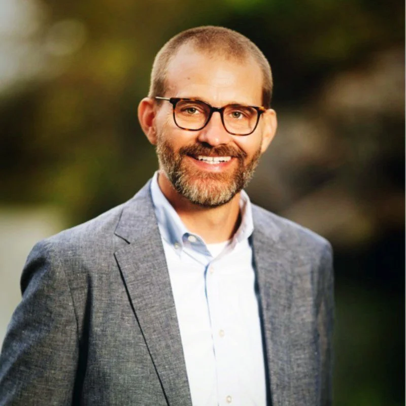 A man with glasses, a beard, and a smile, wearing a light blue shirt and a gray blazer, outdoors with a blurred natural background.