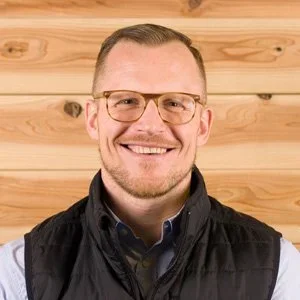 A smiling man with short hair, glasses, and a beard, wearing a black vest over a dark shirt, standing in front of a wooden wall.