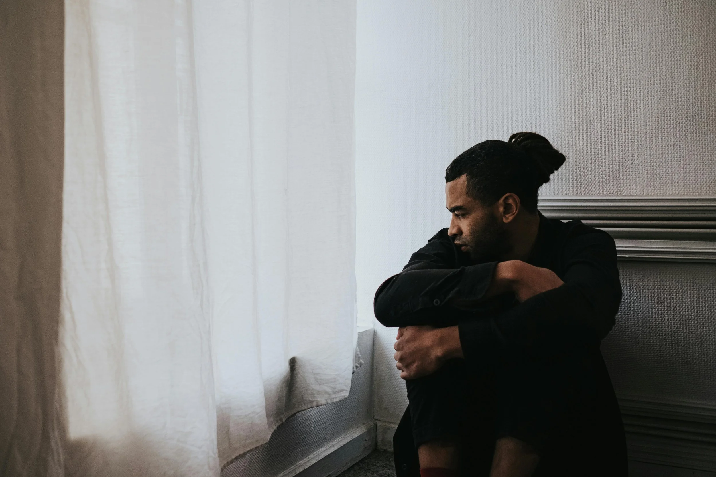 A man with well-groomed black hair in a top knot bun, wearing a black shirt, sitting on the floor with his arms wrapped around his knees, looking down thoughtfully. He is positioned next to a white wall and a partly open white curtain, in a softly lit room.