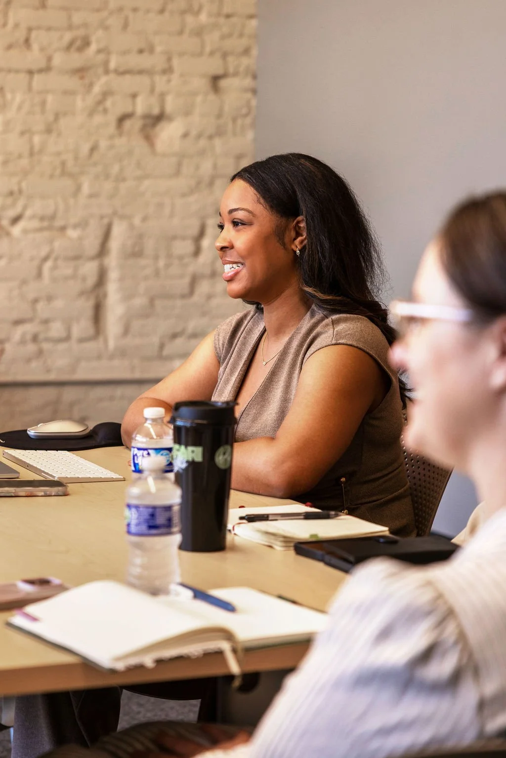 A woman smiling during a meeting in a conference room with notebooks, water bottles, and a coffee tumbler on the table.