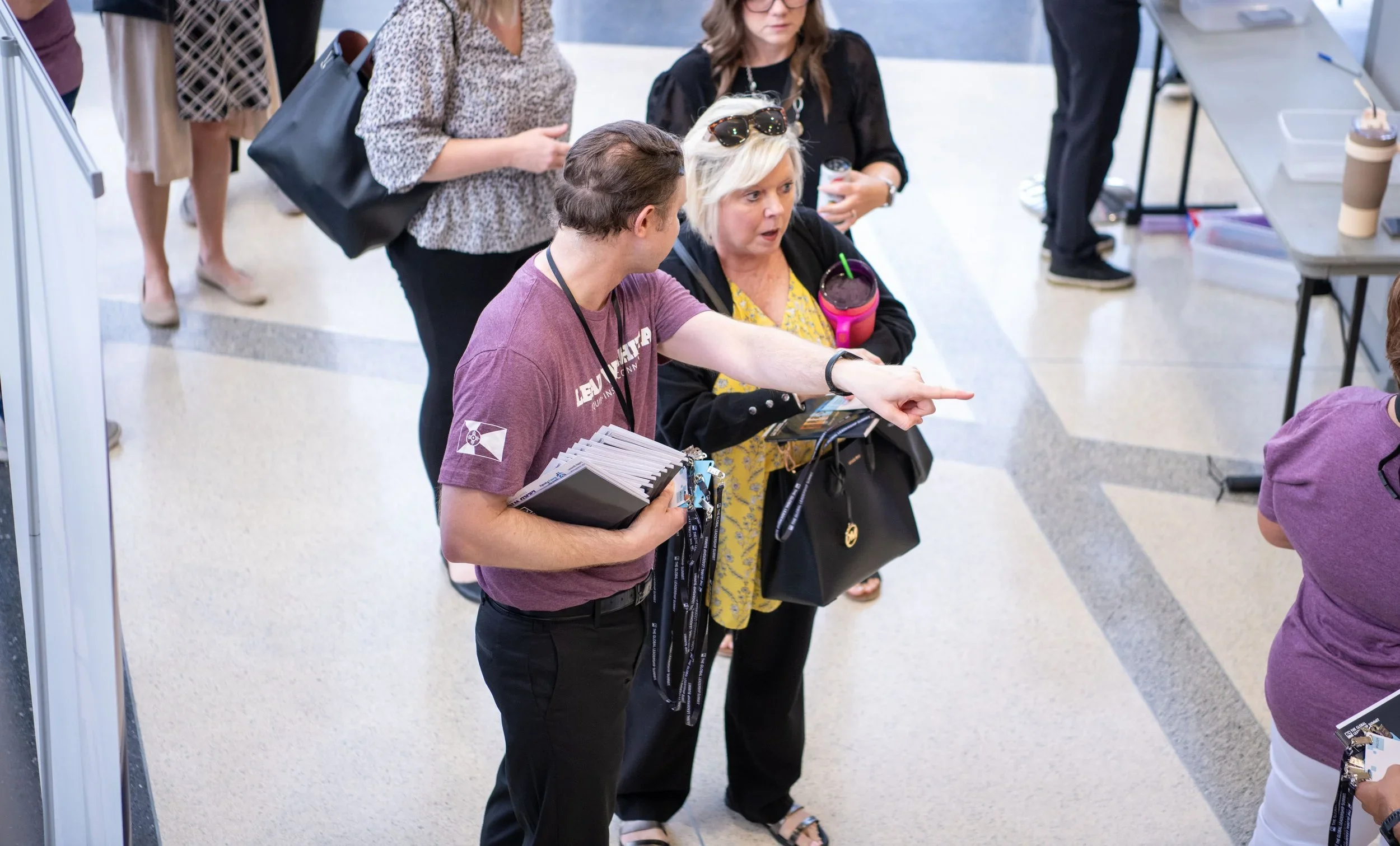 People standing in line at an indoor event, engaging in conversation, with a woman pointing at something and a person holding a clipboard, surrounded by tables and cups.