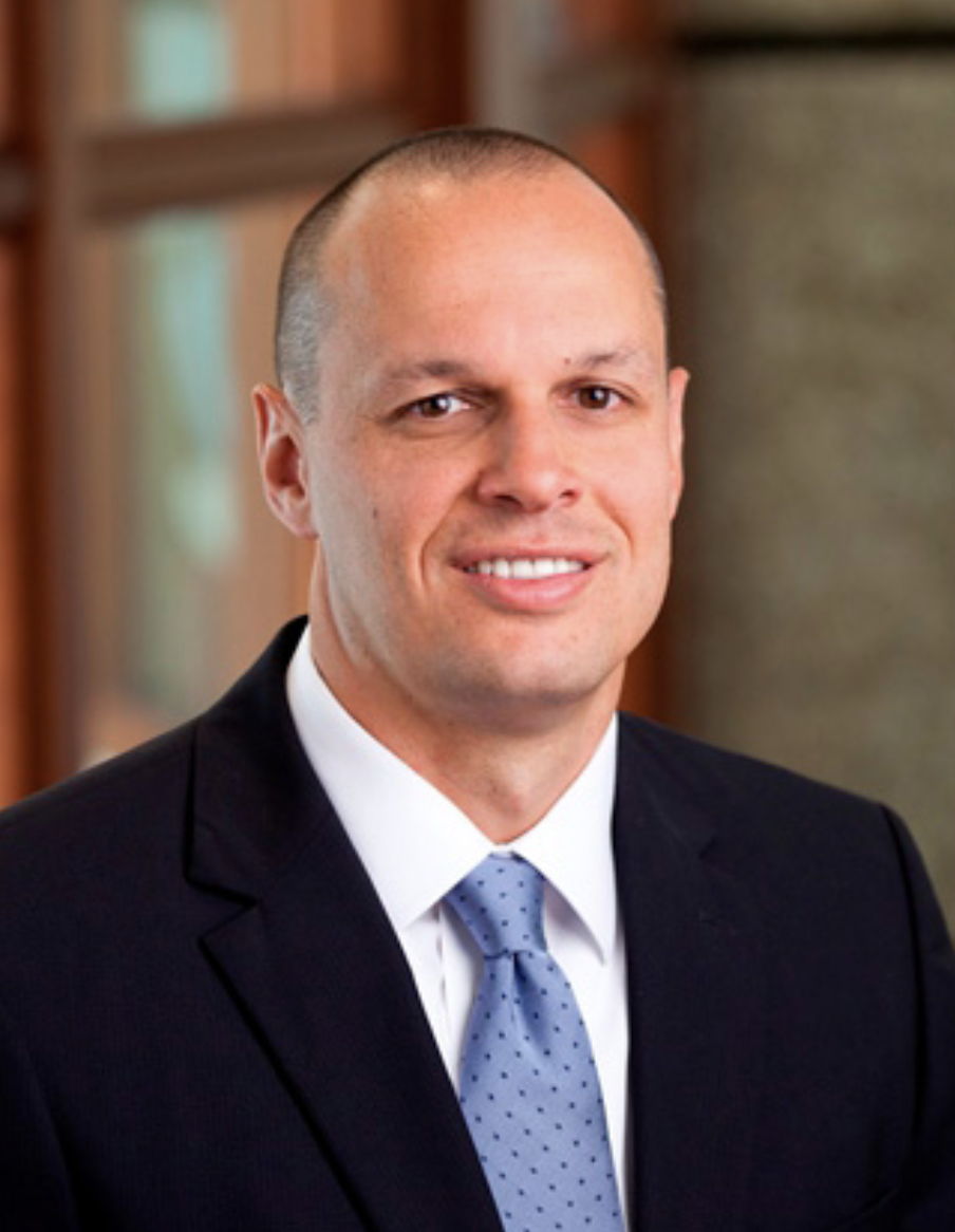 Portrait of a man in a suit and tie, smiling, with a blurred background of wooden furniture.