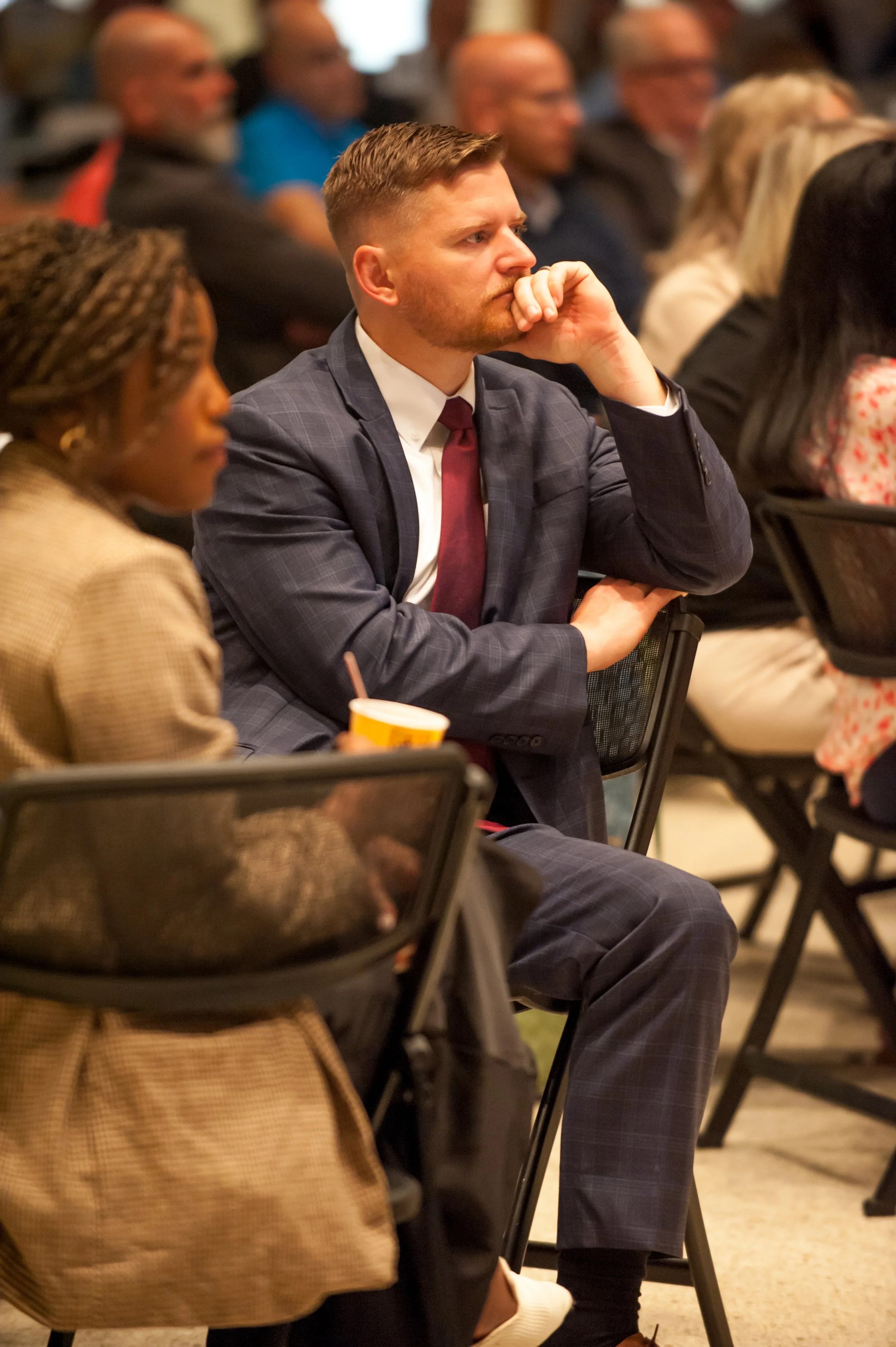 A man in a suit with a maroon tie sitting with his hand on his chin, appearing engaged at a conference or seminar. Other attendees are visible in the background and foreground, seated and listening.