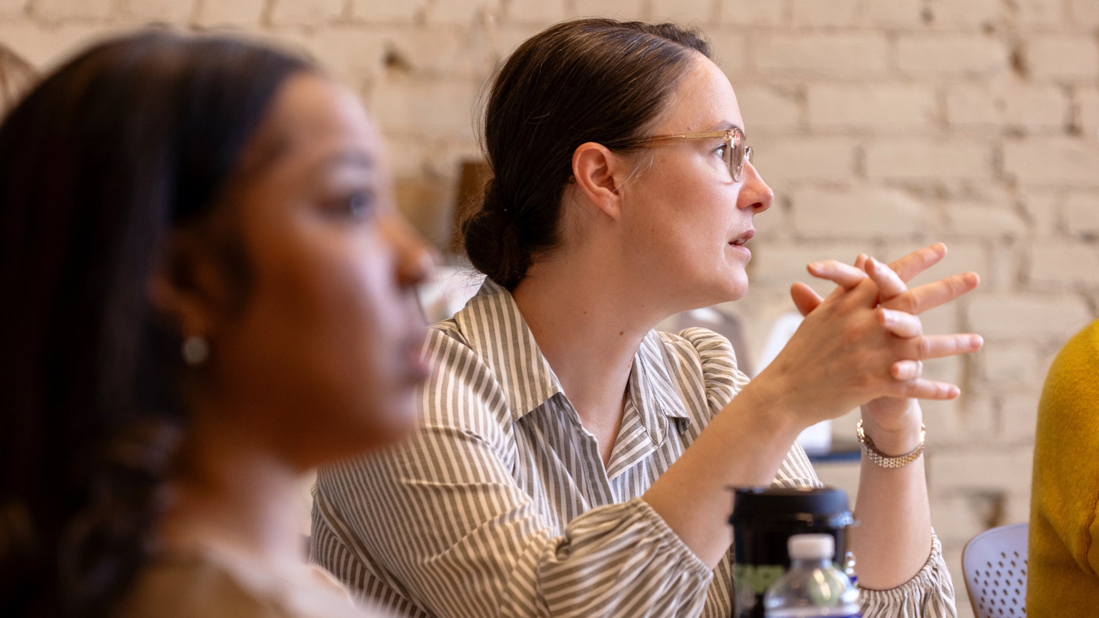 A woman with glasses, wearing a striped shirt, is sitting at a table in a meeting or discussion. She is making a gesture with her hands and appears to be speaking or explaining something. There are other people partially visible around her, and a brick wall is in the background.