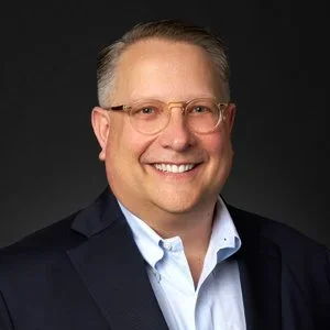 A professional headshot of a smiling man with glasses, wearing a dark suit jacket and a light blue collared shirt against a dark background.