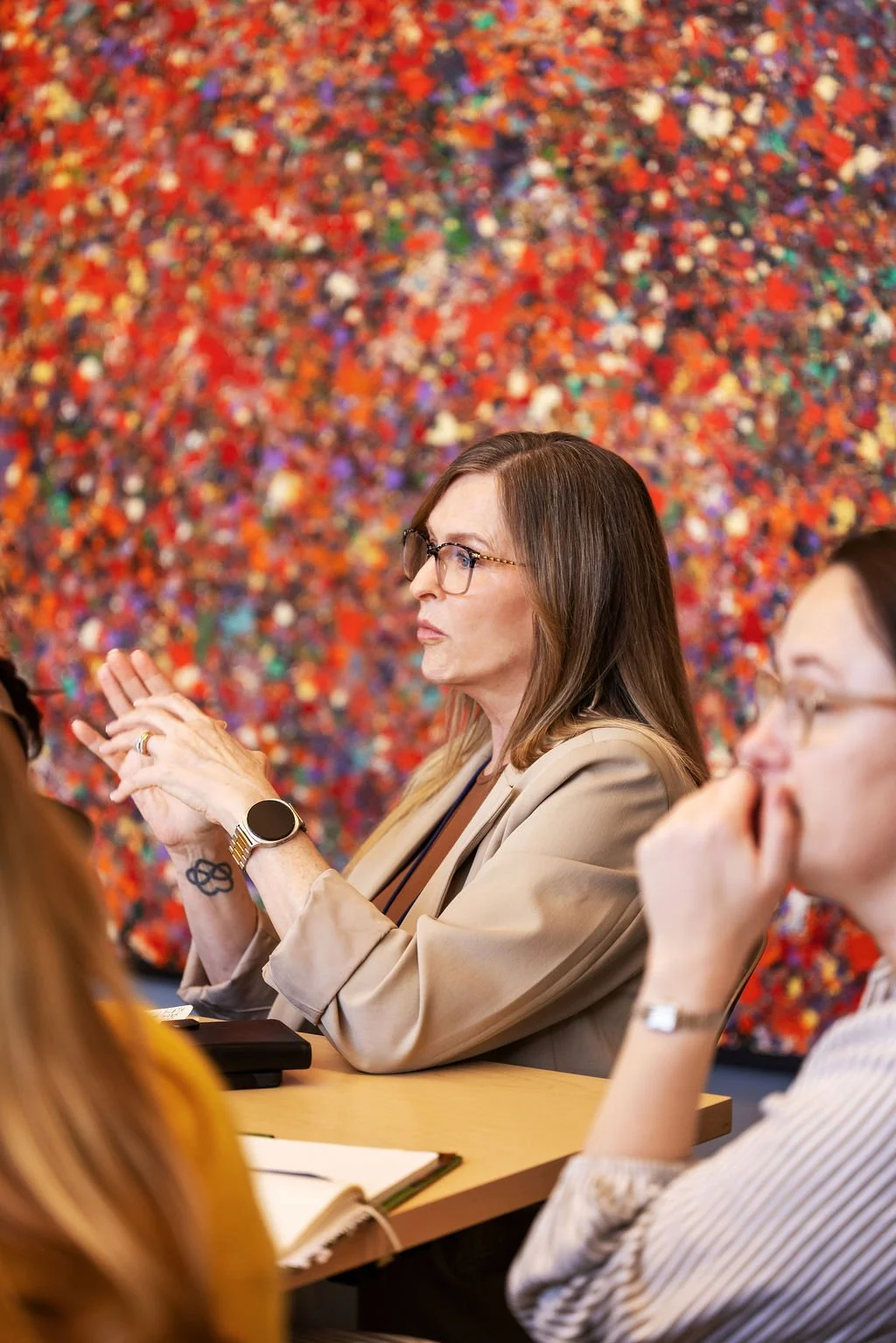 A woman with glasses, wearing a beige blazer, sitting at a table during a meeting, with a colorful abstract art piece in the background.