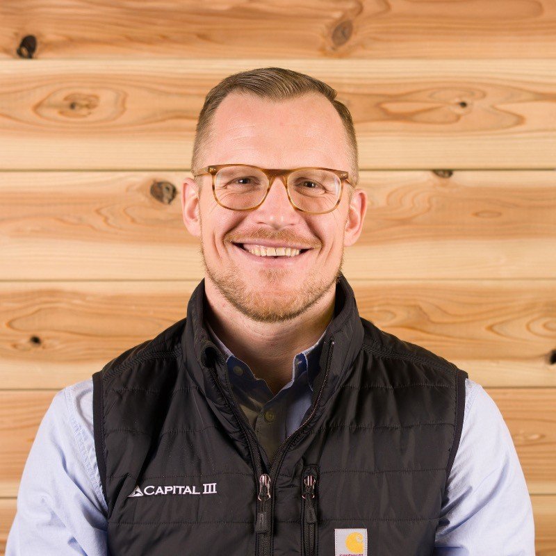 A smiling man with short hair, glasses, and a beard, wearing a black vest over a blue collared shirt, standing in front of a wooden wall.