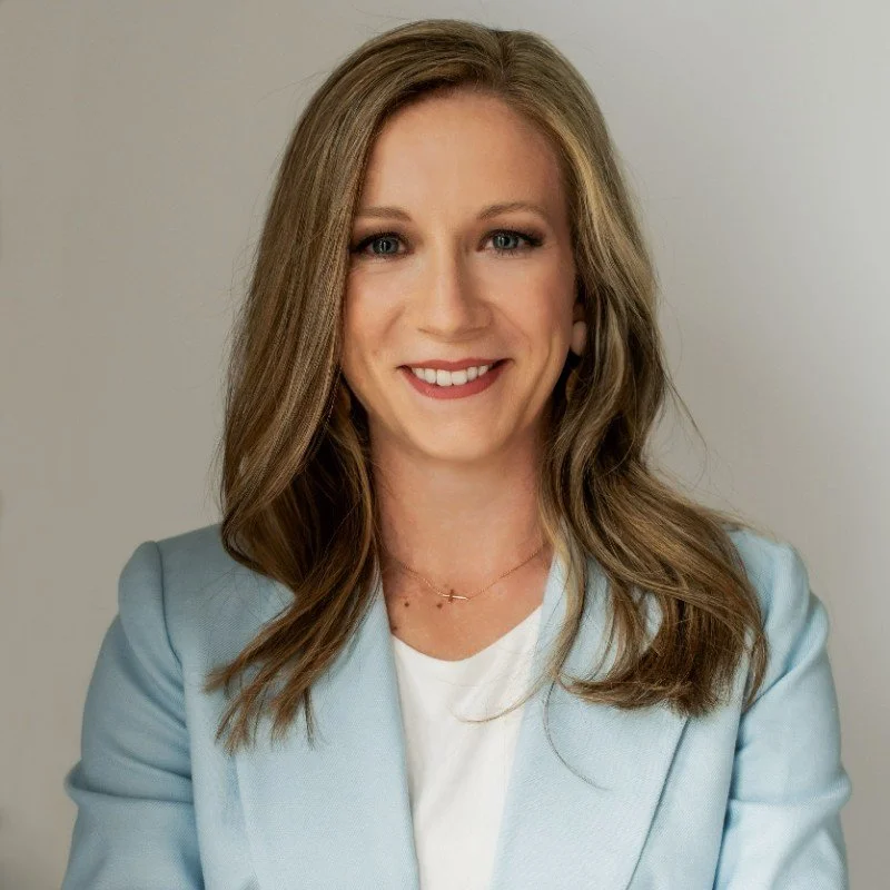 Professional woman with long, wavy, light brown hair, smiling, wearing a light blue blazer and white top, posing against a neutral background.