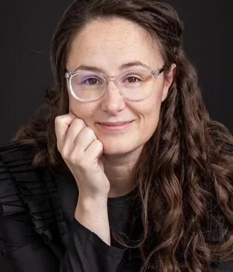 Portrait of a woman with wavy brown hair wearing glasses, resting her chin on her hand, and smiling lightly.