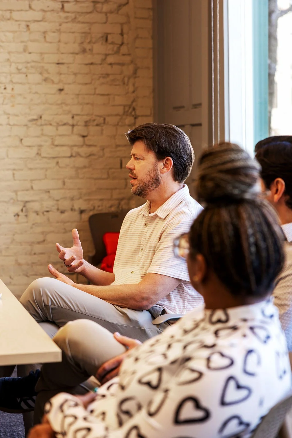 Man in a striped shirt speaking to a group of people in a room with brick wall and large window