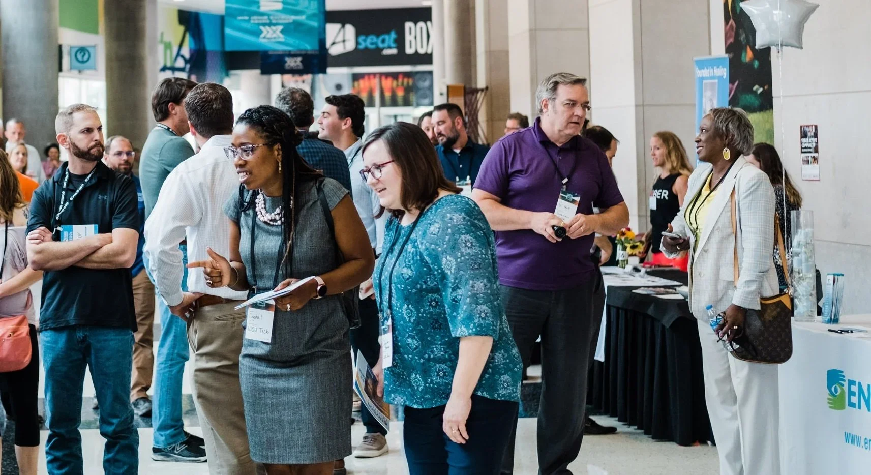 People attending a conference or event in a large indoor space, engaging in conversations, with display booths and banners in the background.