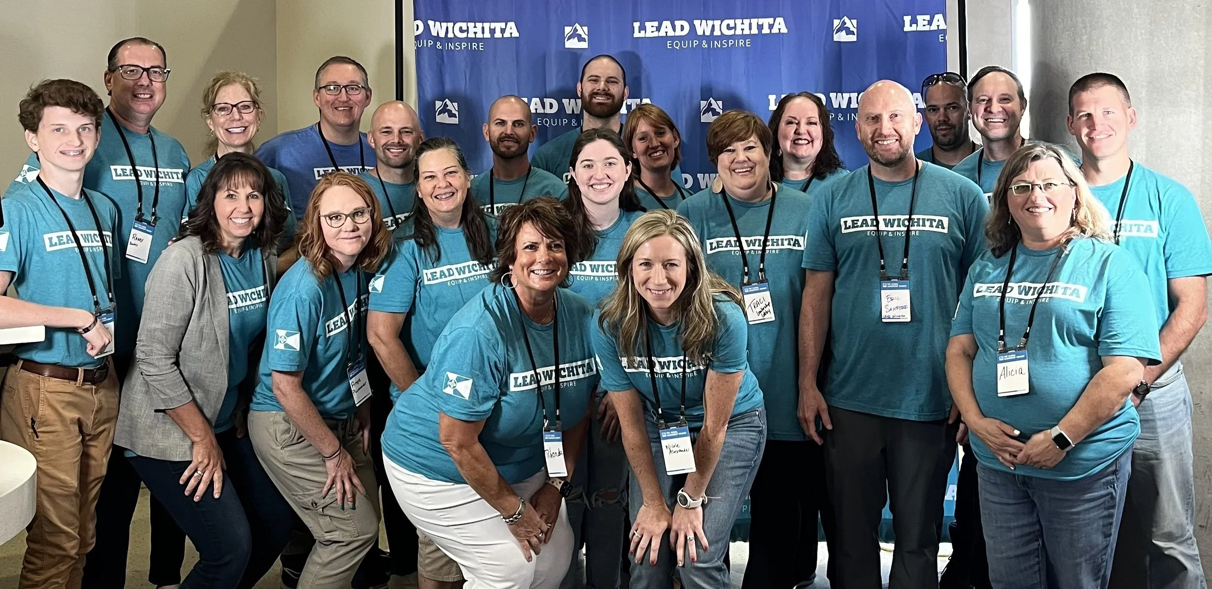 Group of people wearing matching turquoise t-shirts with 'Lead Wichita' logo, posing together at an event in front of a blue backdrop with the same logo.