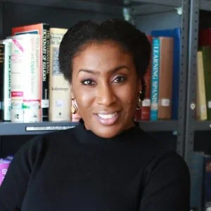Portrait of a woman with short curly hair wearing a black top, standing in front of a bookshelf filled with books.
