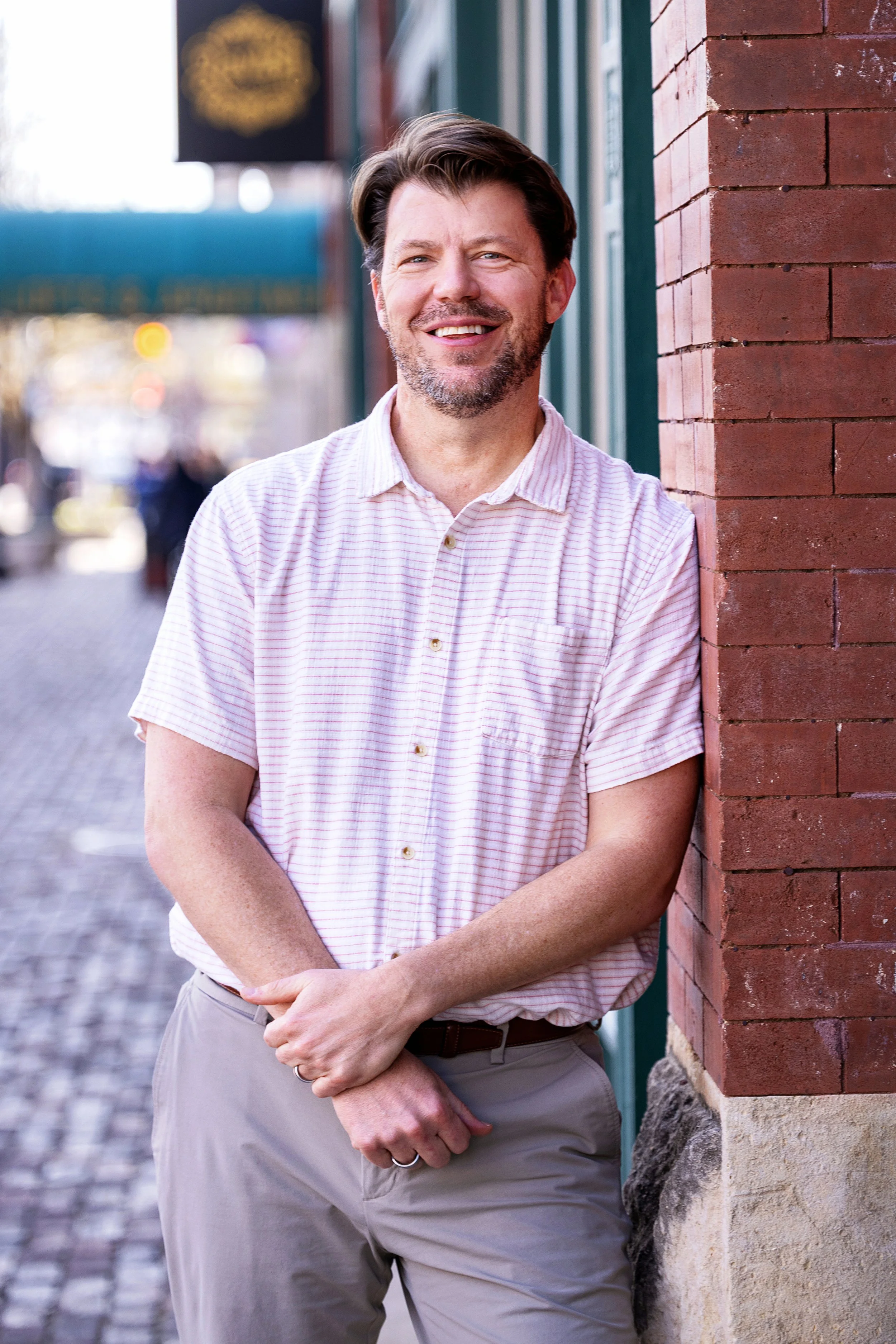 A man standing outdoors next to a brick wall with a blurred street background, smiling, wearing a light-colored, striped, short-sleeved button-up shirt and beige pants.