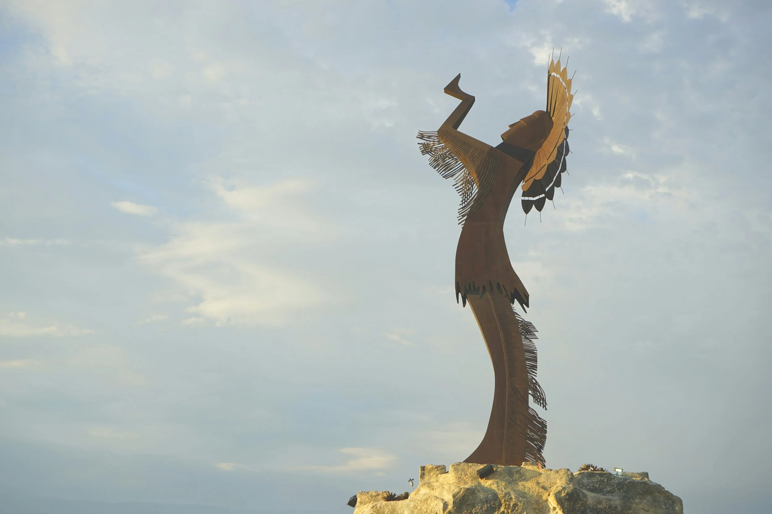 A large metal sculpture resembling a stylized bird with outstretched wings and a long neck, situated on a rocky hilltop with a cloudy sky in the background.