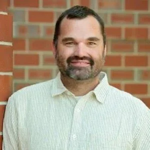 A man with dark hair and a beard smiling, wearing a light-colored collared shirt, standing in front of a brick wall.
