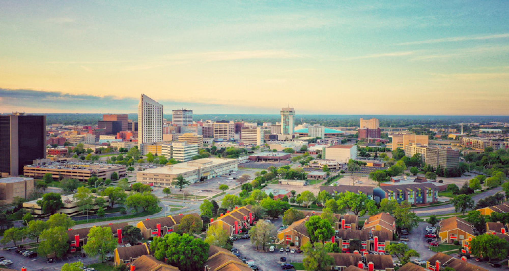 A city skyline during daylight with various tall buildings, a tower, and lush green trees in the foreground. Residential neighborhoods with red-roofed houses are also visible.