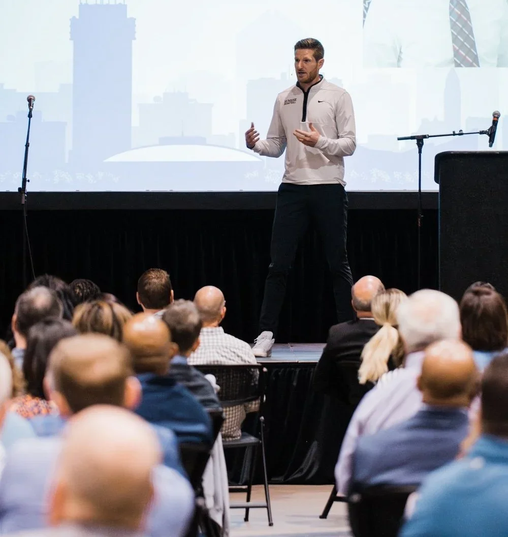 Man giving a presentation on stage in front of an audience with a cityscape background projected behind him.