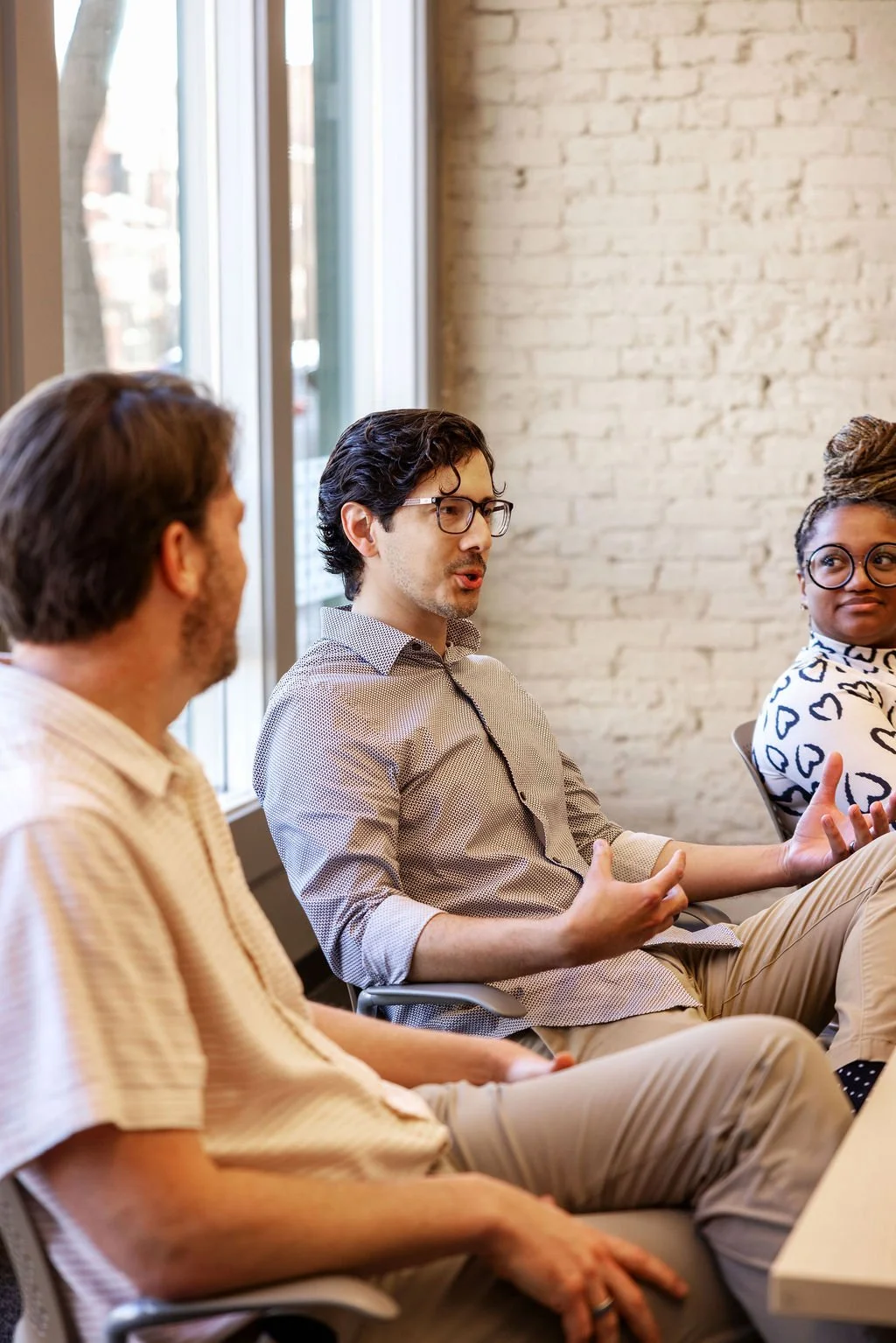 Three people engaged in a discussion in a modern office with exposed brick wall and large windows.