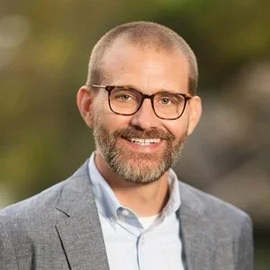 A man with short hair, beard, and glasses wearing a light gray suit jacket over a white shirt, smiling outdoors with a blurred natural background.
