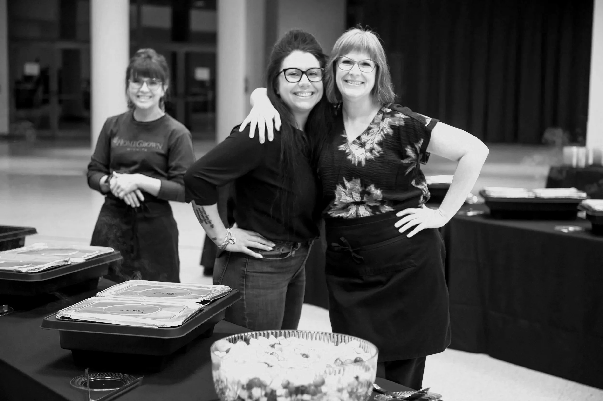 Three women smiling at the camera, standing behind a table with trays of food and snacks in a room with dark curtains and a stage in the background.