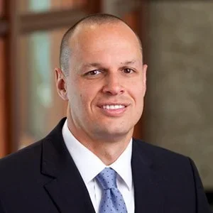 A man in a black suit, white shirt, and blue tie smiling indoors with a blurred background.