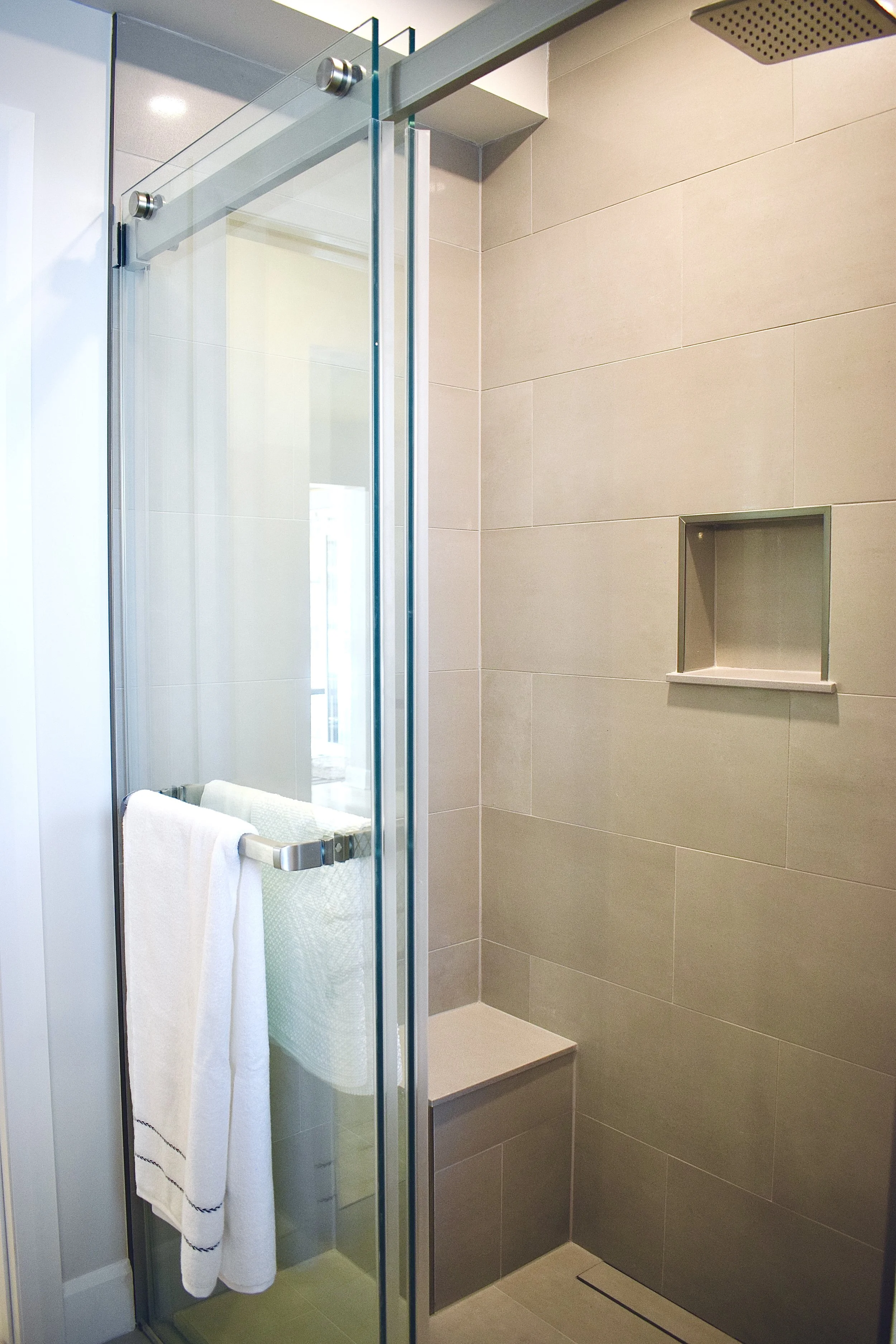 View of a modern shower with glass door, beige tiled walls, a built-in niche, and a rainfall showerhead.