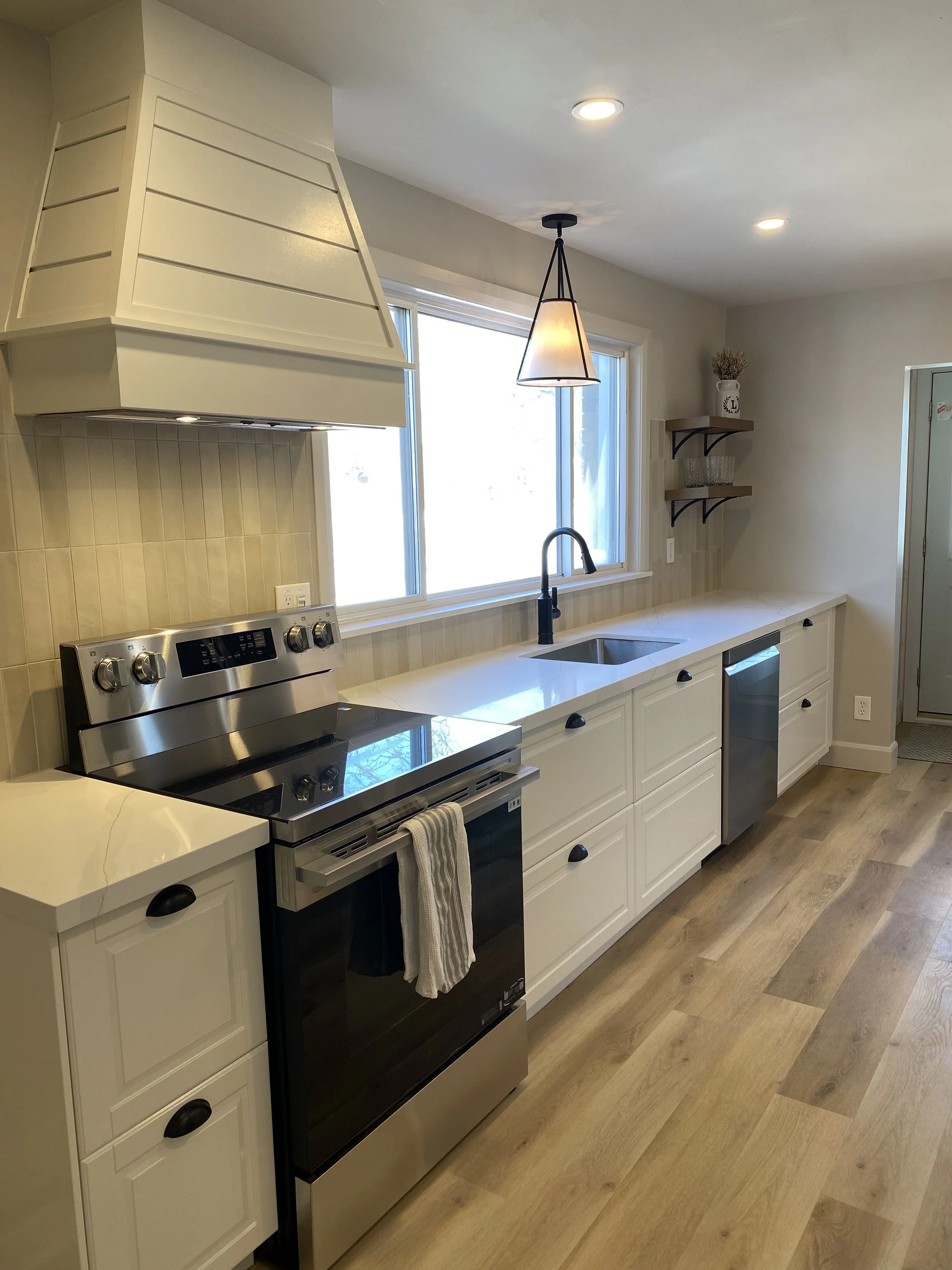 A modern kitchen with white cabinets, a black dishwasher, a stainless steel stove, a black faucet, a large window, and two ceiling lights.