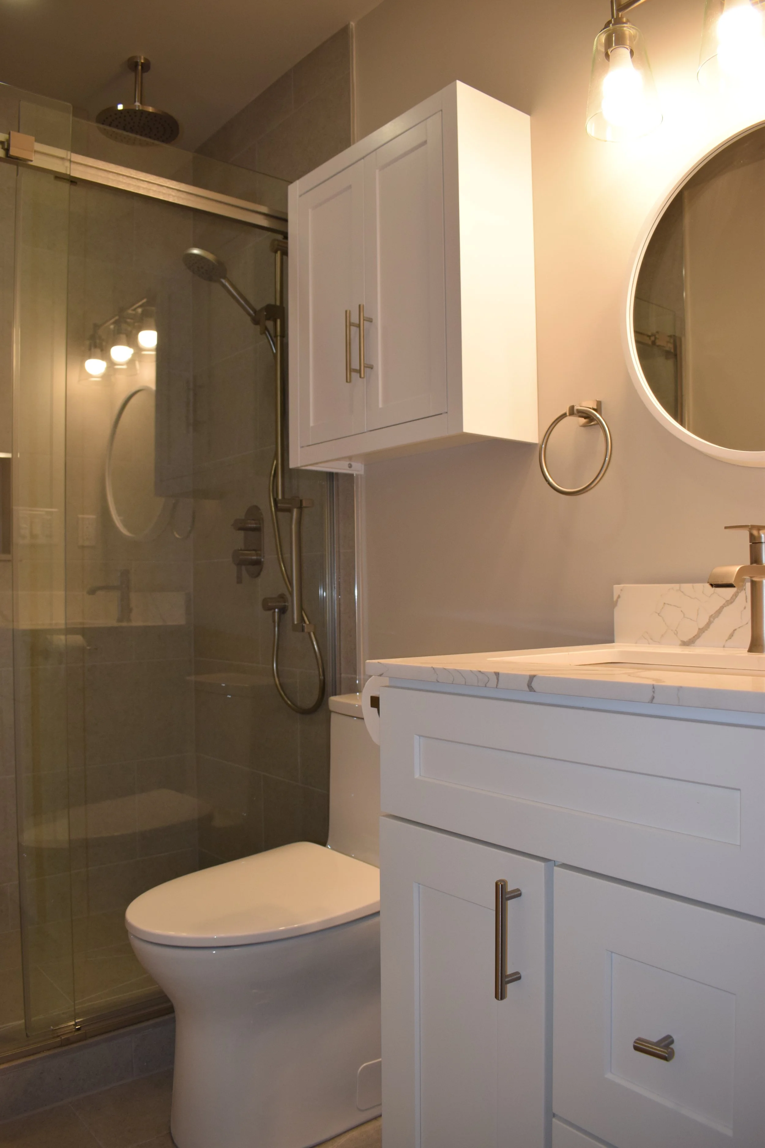A modern bathroom with a walk-in shower, white vanity with drawers, a large oval mirror, and a toilet. The shower has a glass door and two showerheads, with a neutral beige and gray color scheme.