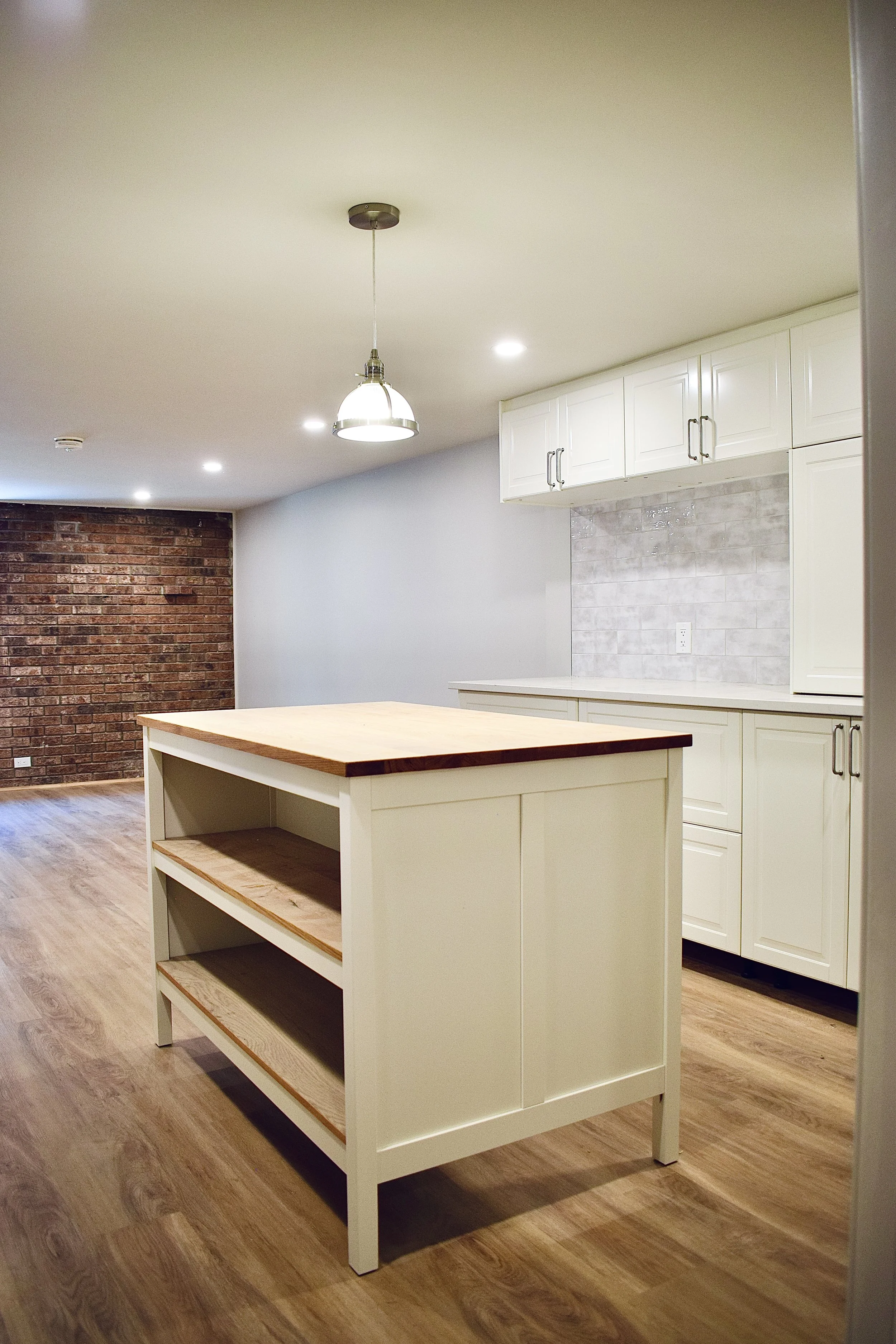 Modern kitchen with white cabinets, a central island with open shelves, a brick accent wall, and wooden flooring, illuminated by ceiling lights and a hanging pendant light.