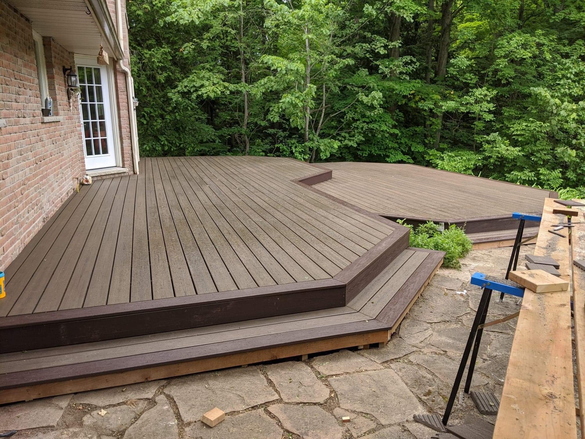 Backyard with a new wooden deck attached to a brick house, surrounded by dense green trees.