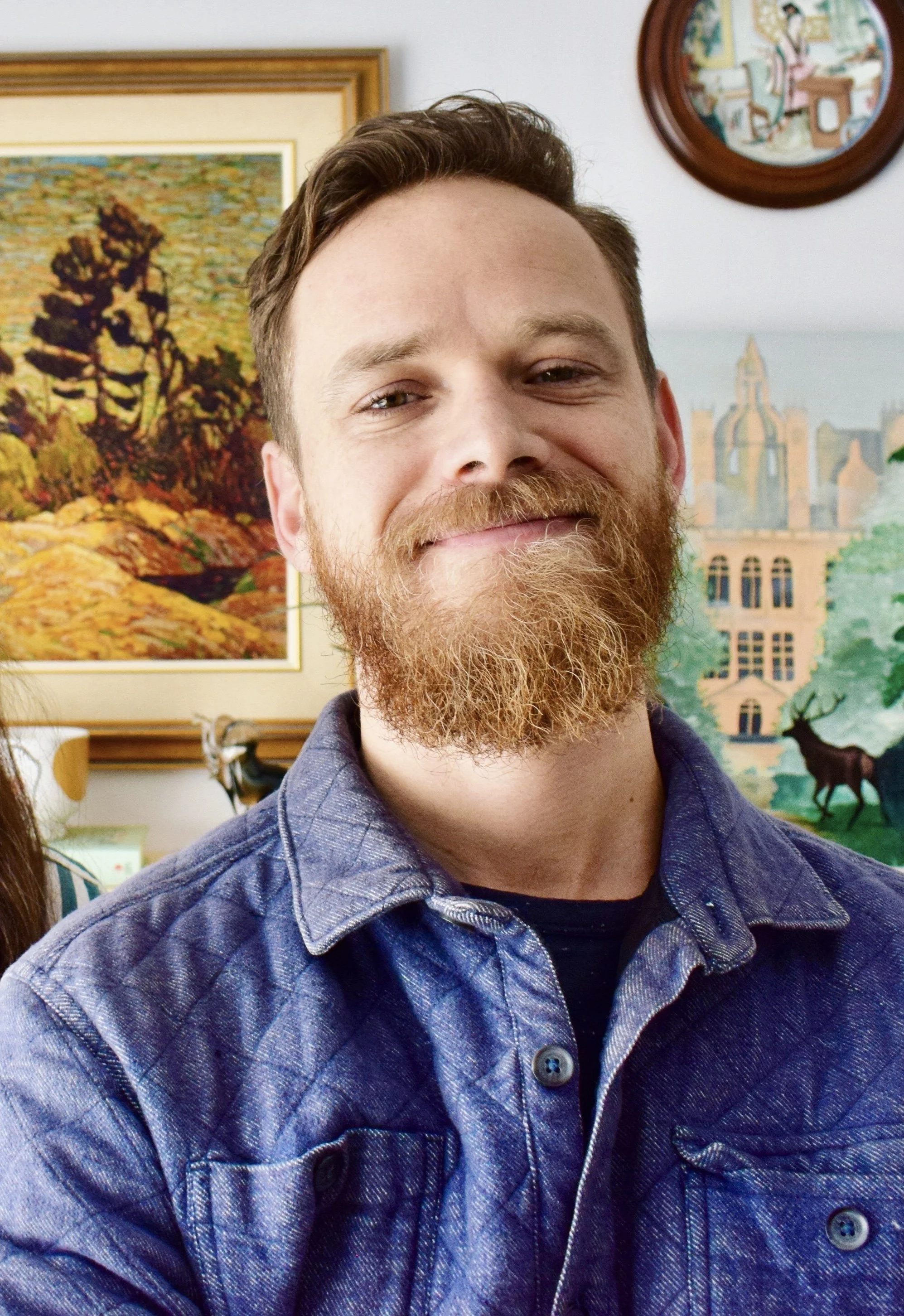 Close-up of a smiling man with a beard wearing a blue denim shirt. The background shows colorful paintings and a circular artwork hanging on the wall.