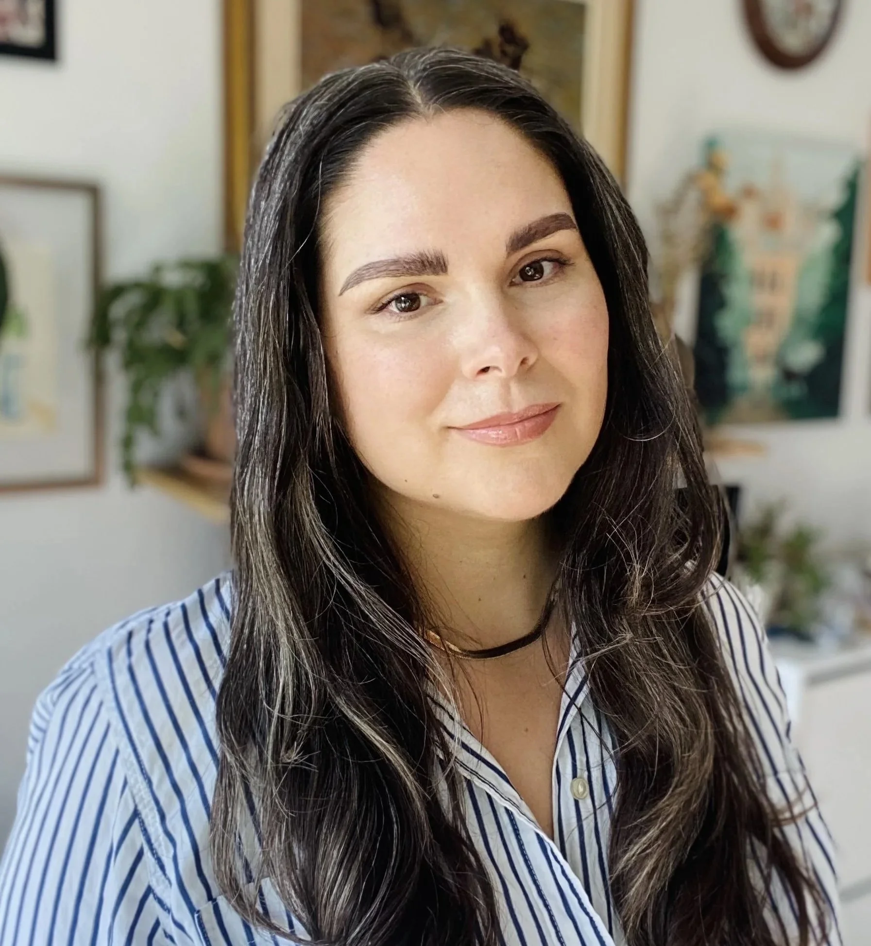 Close-up of a woman with long dark hair, wearing a striped shirt, in a room with artwork and a plant in the background.