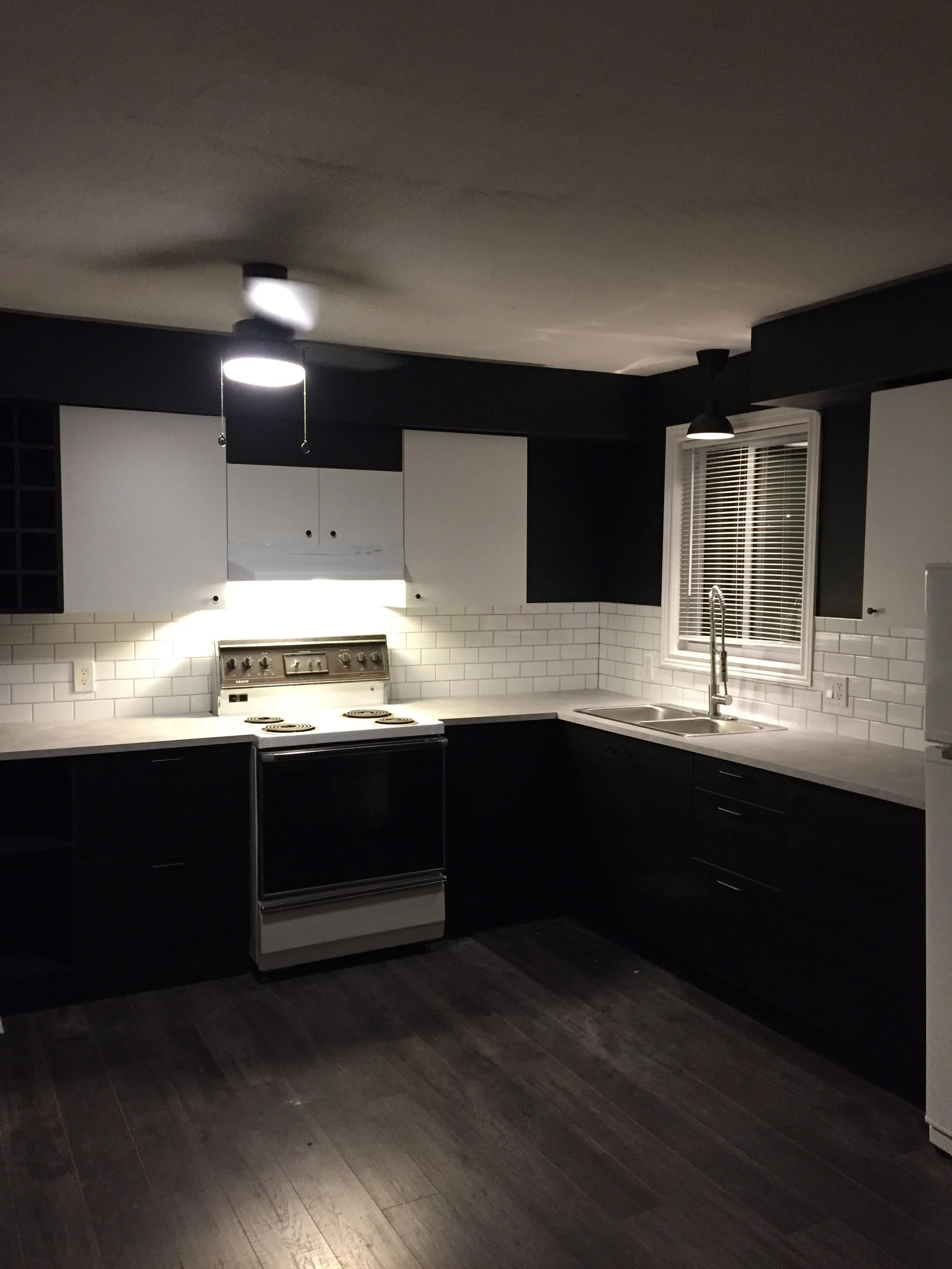 A modern kitchen with black lower cabinets, white upper cabinets, a white subway tile backsplash, a stainless steel stove, and a double sink. There is a window with blinds above the sink, and a ceiling fan with a light.