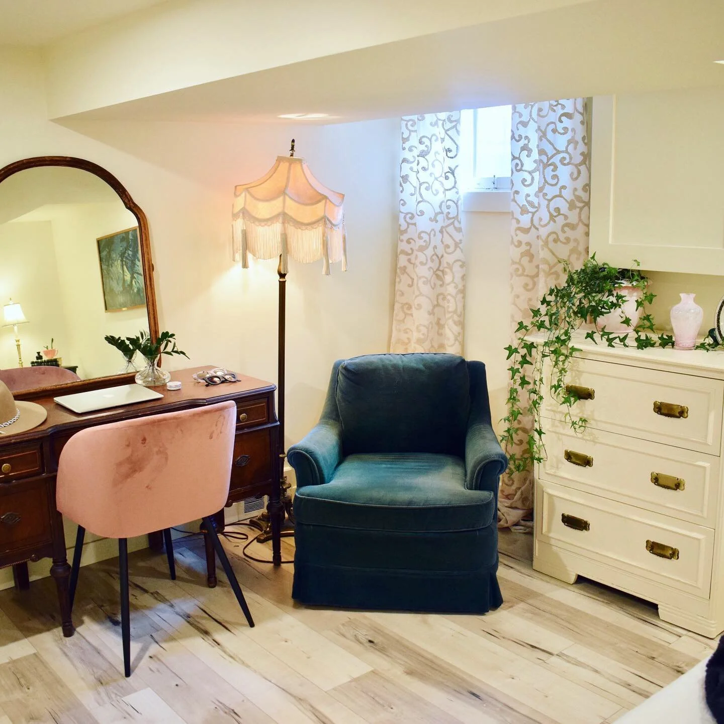 Living room with a vintage dresser, mirror, pink chair, teal armchair, a window with floral curtains, and a white dresser with plants and vases.