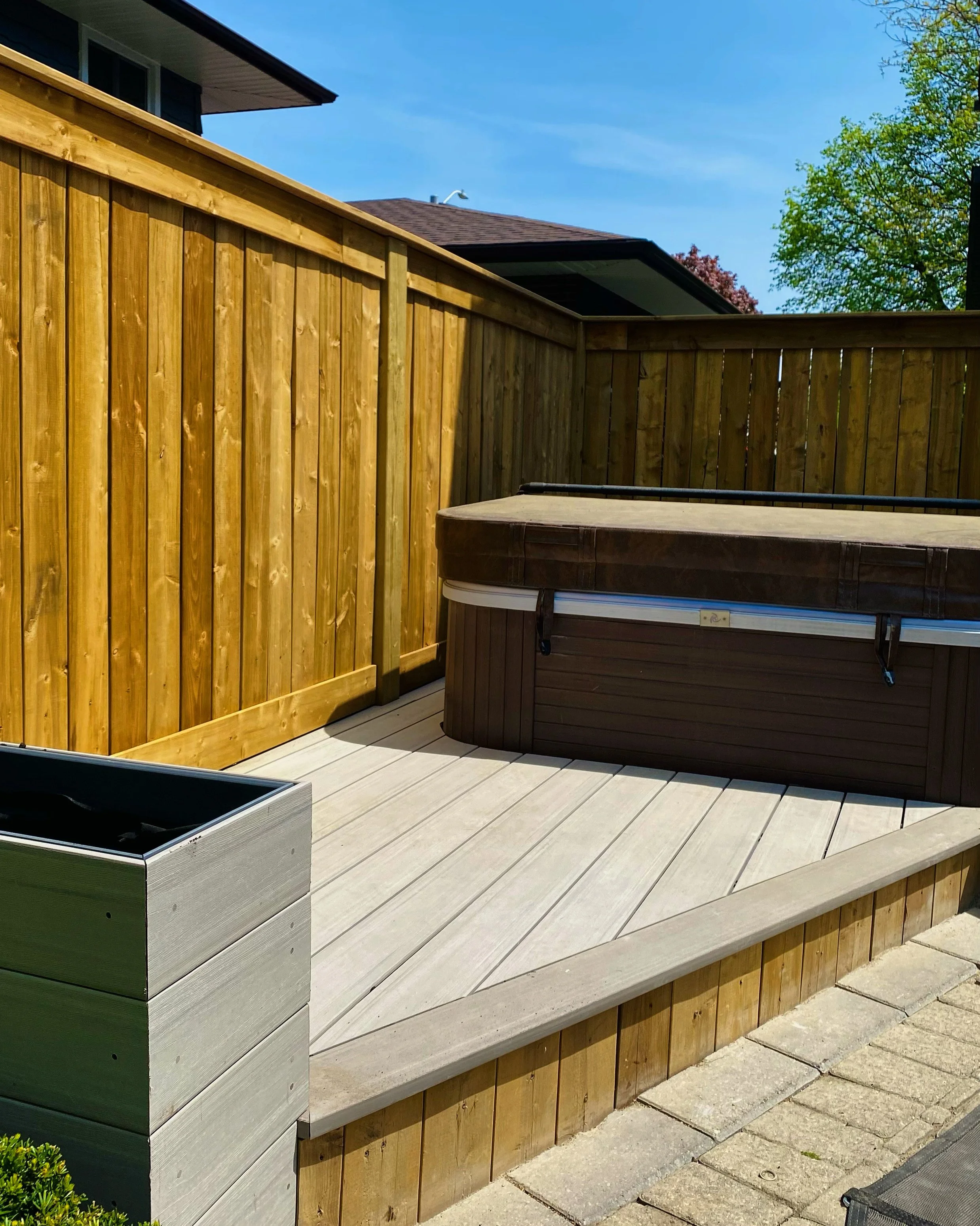 A backyard scene with a wooden privacy fence, a hot tub, and a patio with light-colored tiles. In the background, there is a house with a sloped roof, trees, and a clear blue sky.