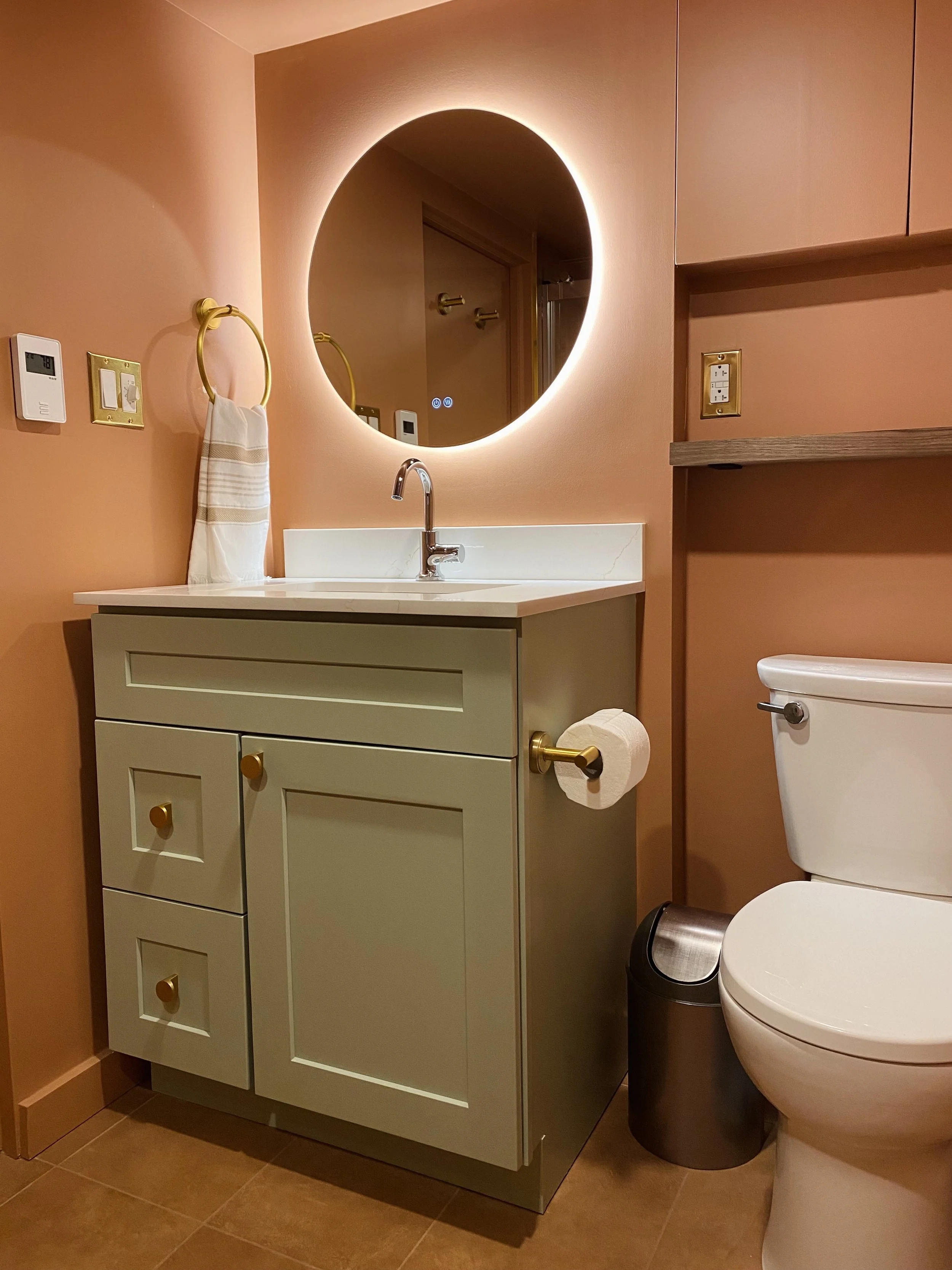 A bathroom with peach-colored walls, a white vanity with gold knobs, a round mirror with backlighting, a towel on a gold ring, a toilet with a black trash bin, and brown floor tiles.