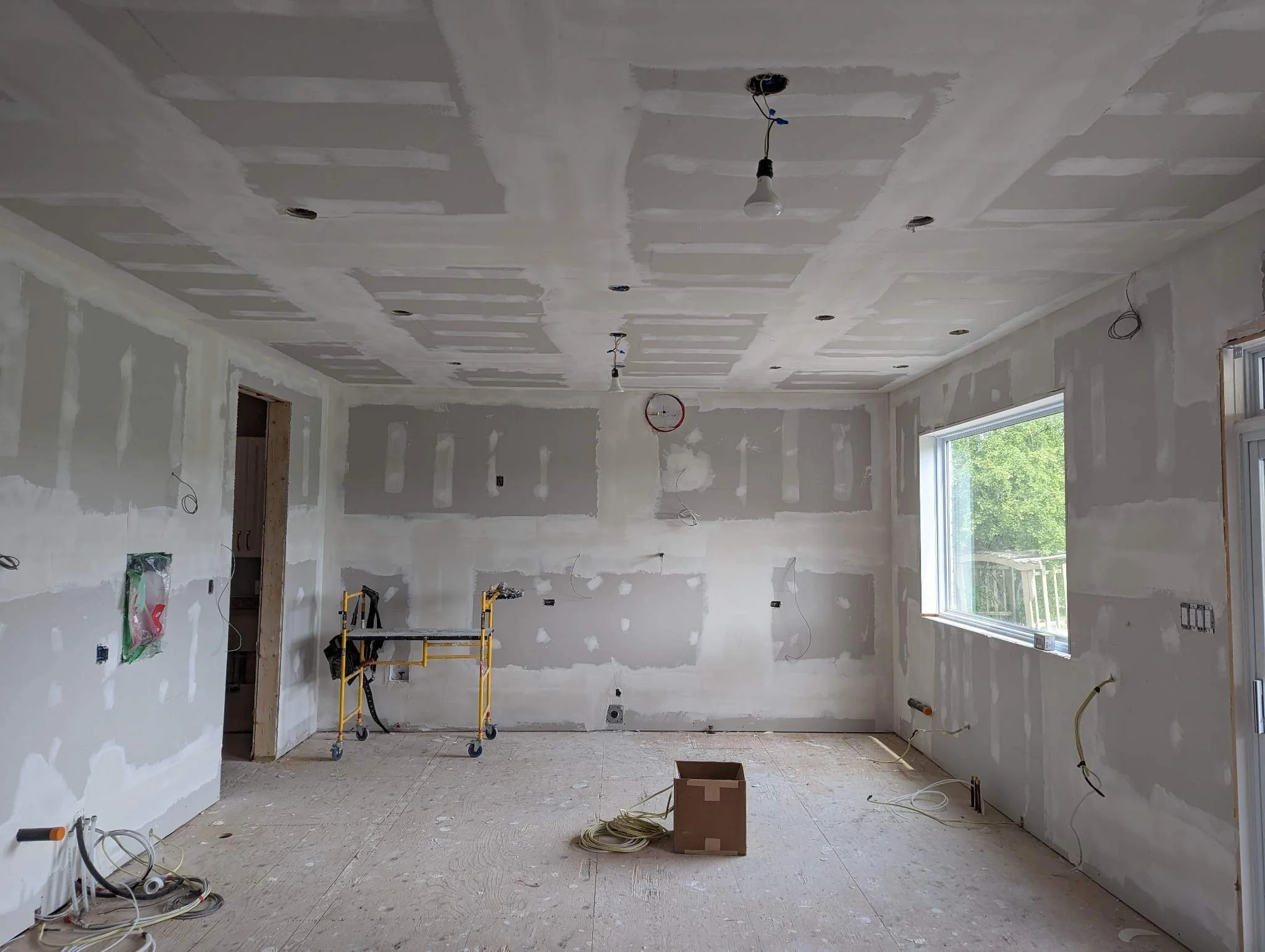 An unfinished interior room under construction with drywall walls and ceiling, exposed lighting fixtures, electrical wires, a small scaffolding, and a large window showing green trees outside.