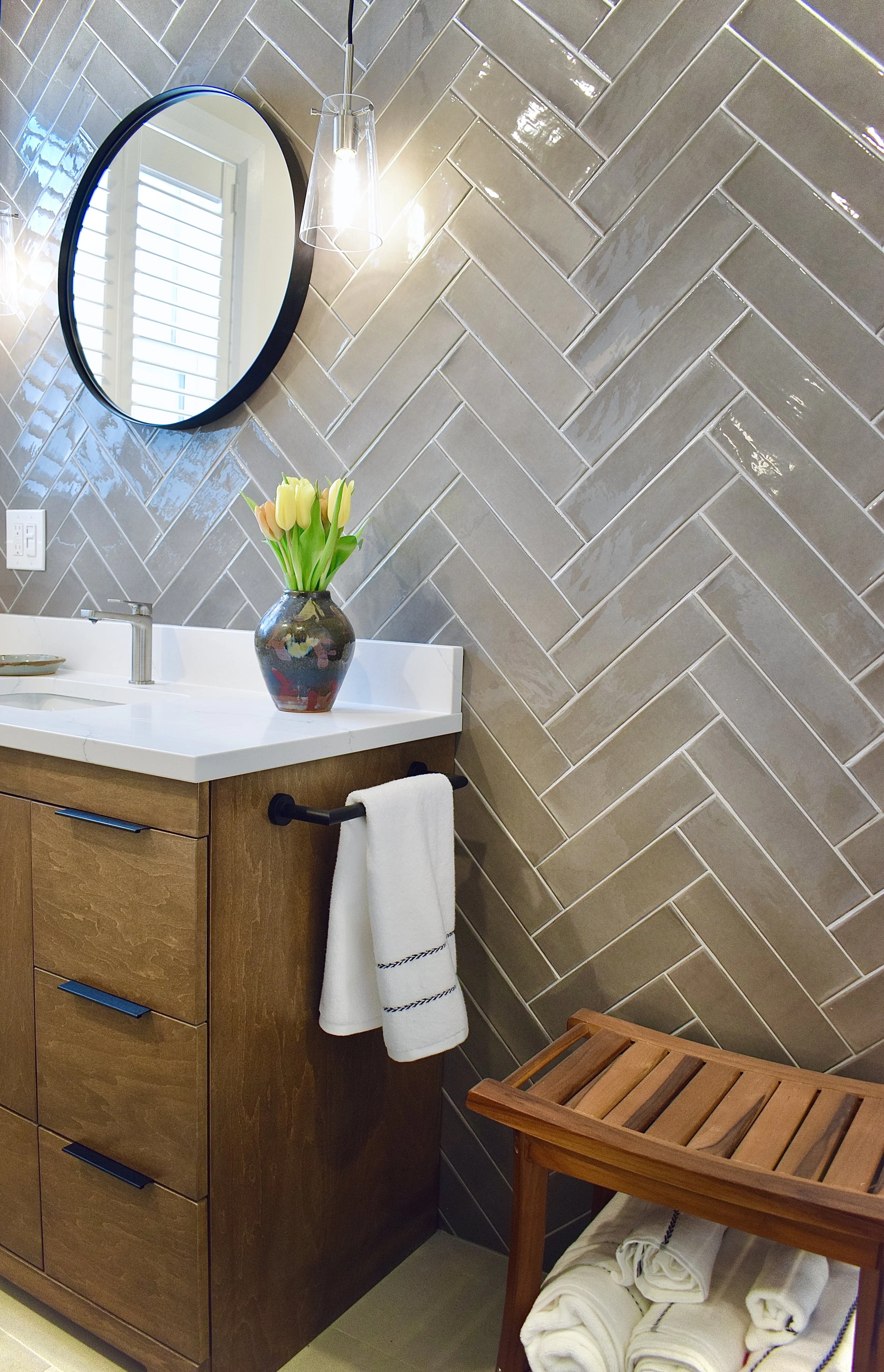 A bathroom with beige tile wall in a herringbone pattern, a white sink with a wooden cabinet below, a black framed round mirror, a glass light fixture, a vase with tulips on the sink, and a wooden bench with rolled towels underneath.